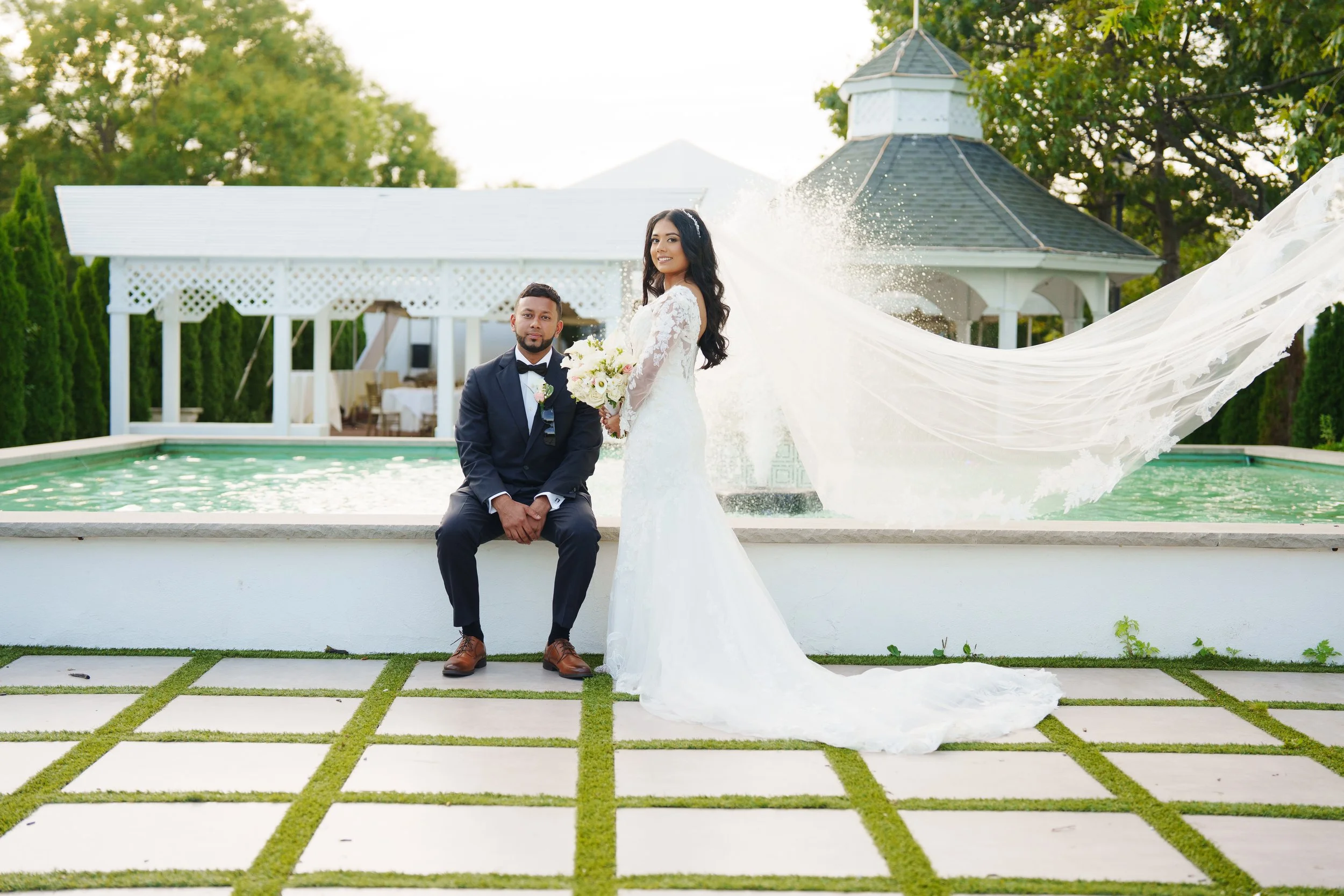 New York City Wedding Videographer & Photographer Dane Nelson Photo of Bride in a long white wedding dress holding a bouquet, standing next to a groom in a black tuxedo, at Worlds Fair Marina wedding venue with a fountain and gazebo.