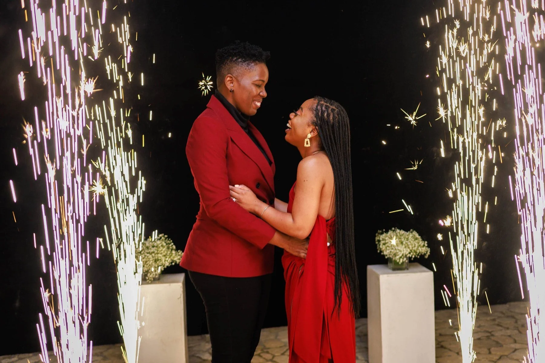 Engaged couple in red embracing and smiling at each other between tall columns of sparks during their engagement session in Jamaica
