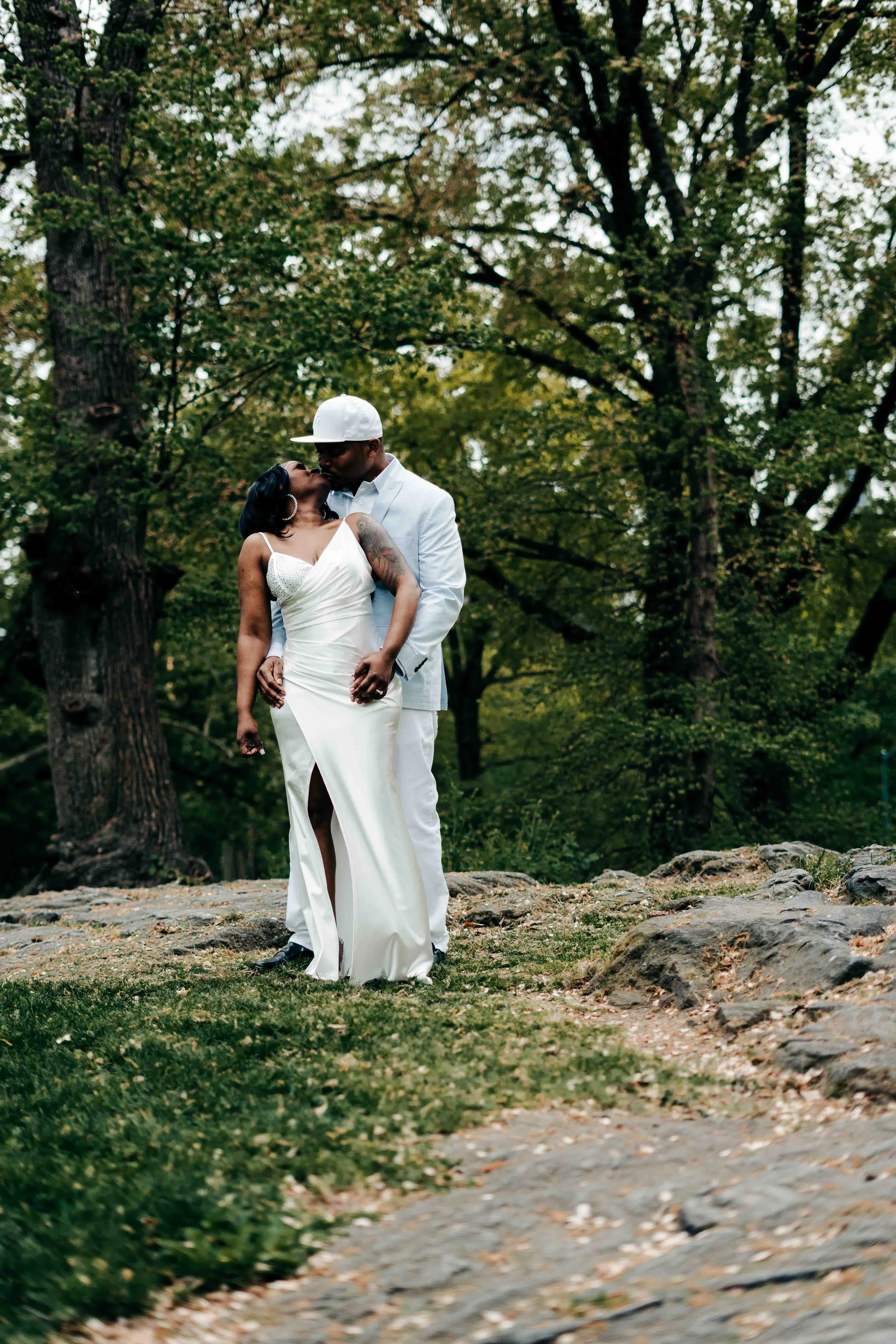 A couple embracing and kissing outdoors; woman in white dress, man in white attire with cap, surrounded by trees in central park