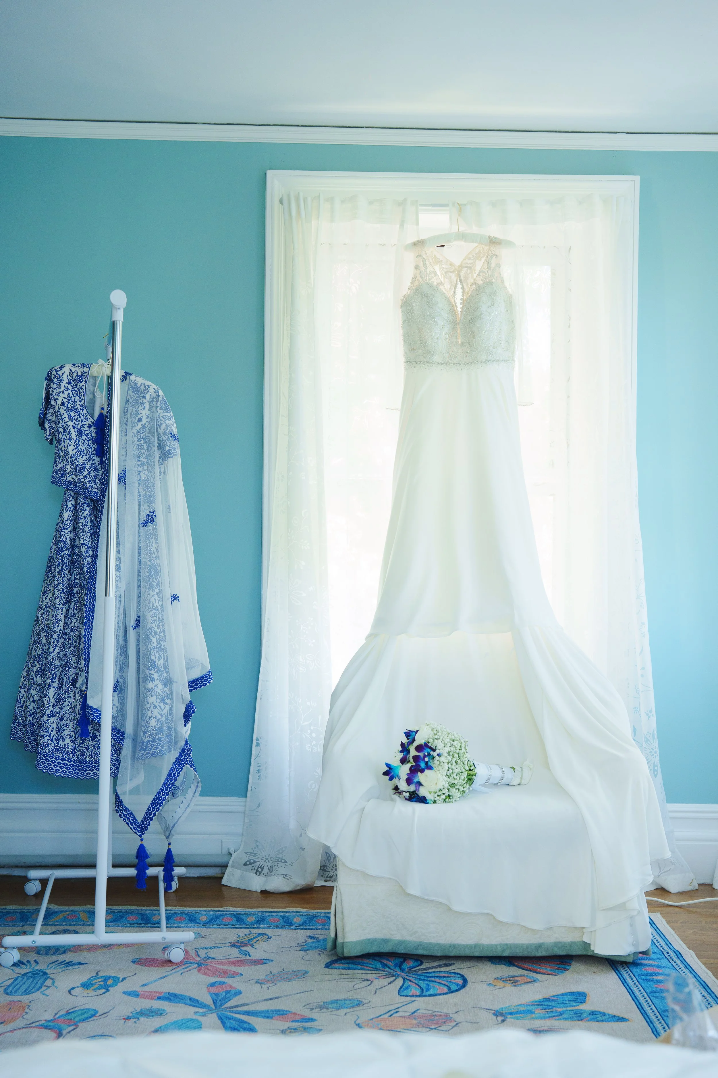 Wedding dress hanging in teal window with Indian lehenga on rack at Arrow Park wedding getting ready suite Monroe NY