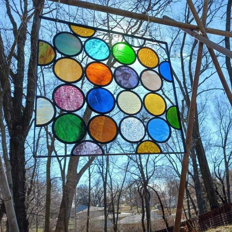 Custom, handmade stained glass art. Square metal piece with multiple different colors of glass soldered into it, hanging outside with sun and trees in background. Created by Artistained Glass.