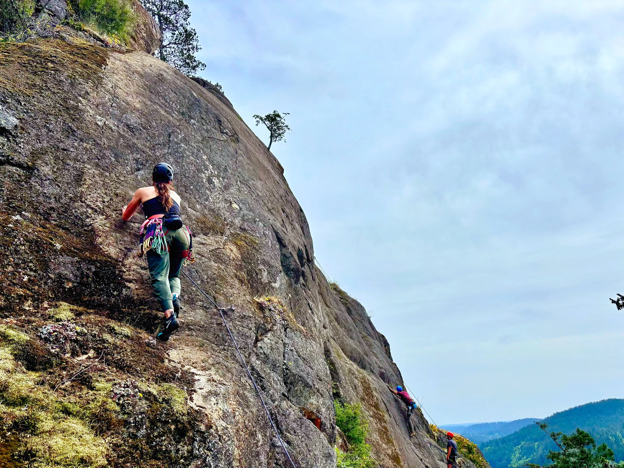 Woman rock climbing outdoors with helmet, climbing gear, and athletic clothing, ascending a rock face.