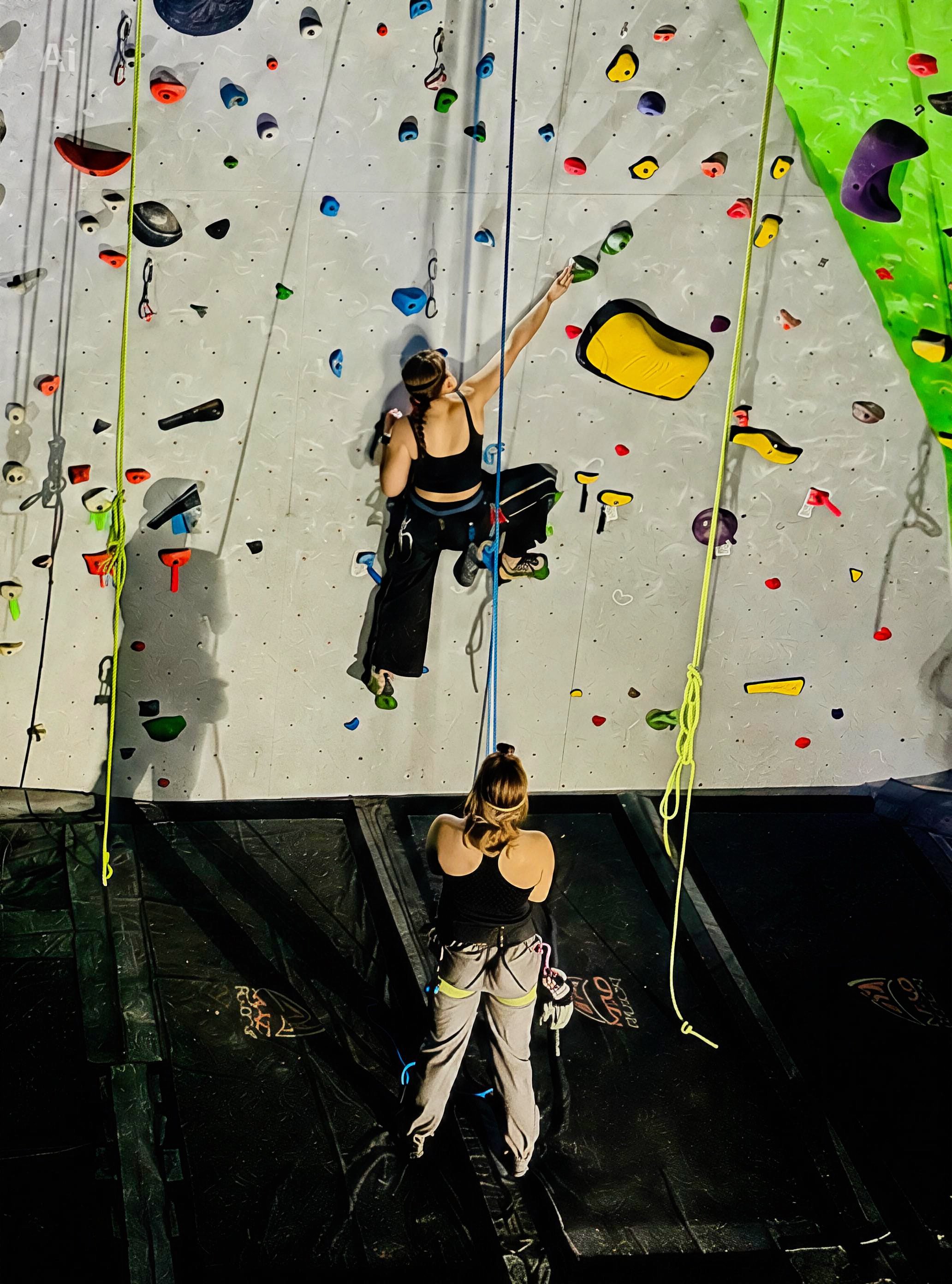 A woman is rock climbing an indoor climbing wall, while a climbing instructor is holding the safety rope and watching her climb.