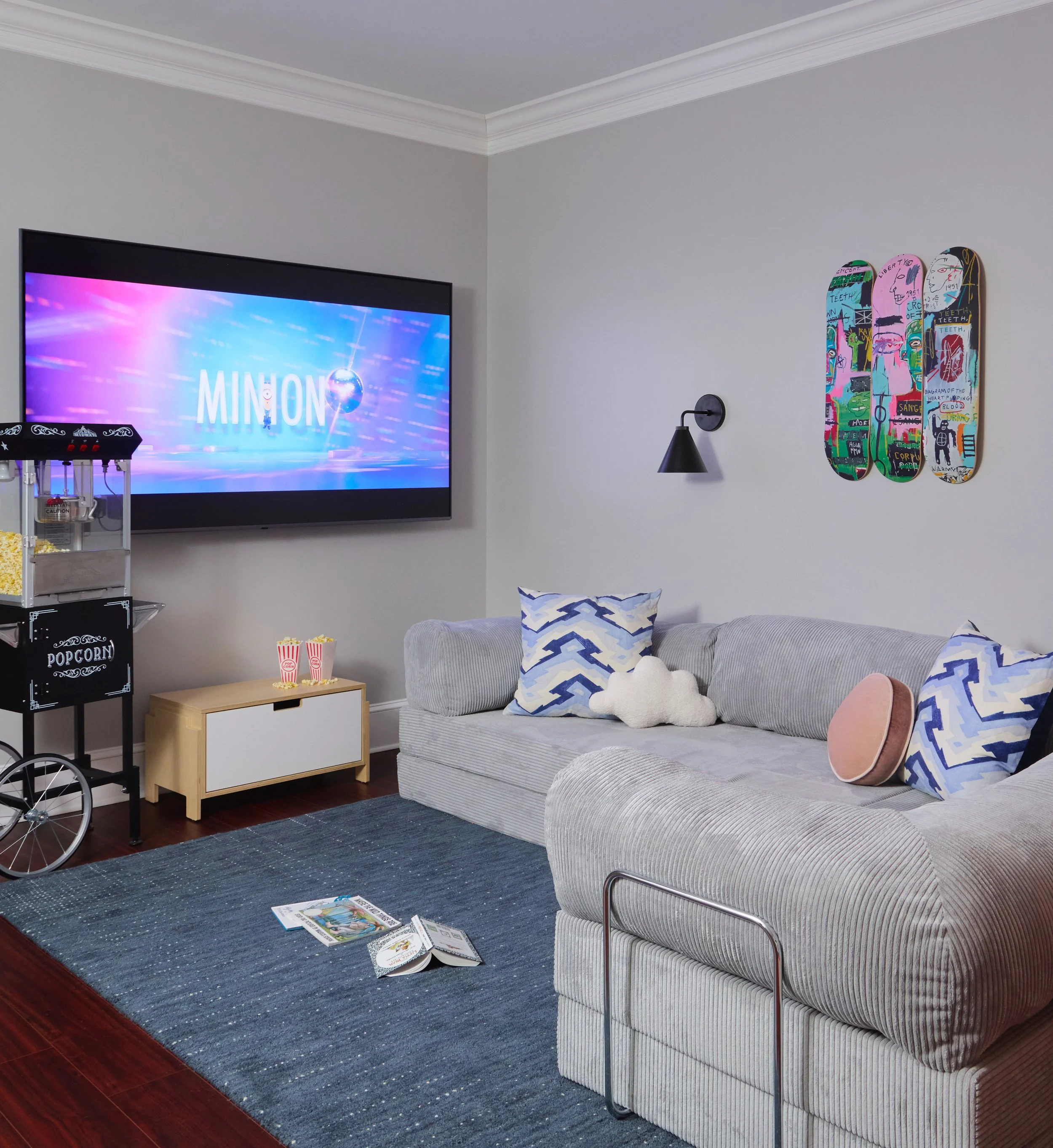 Playroom movie lounge with wall-mounted TV, popcorn machine, grey sectional sofa, and navy area rug