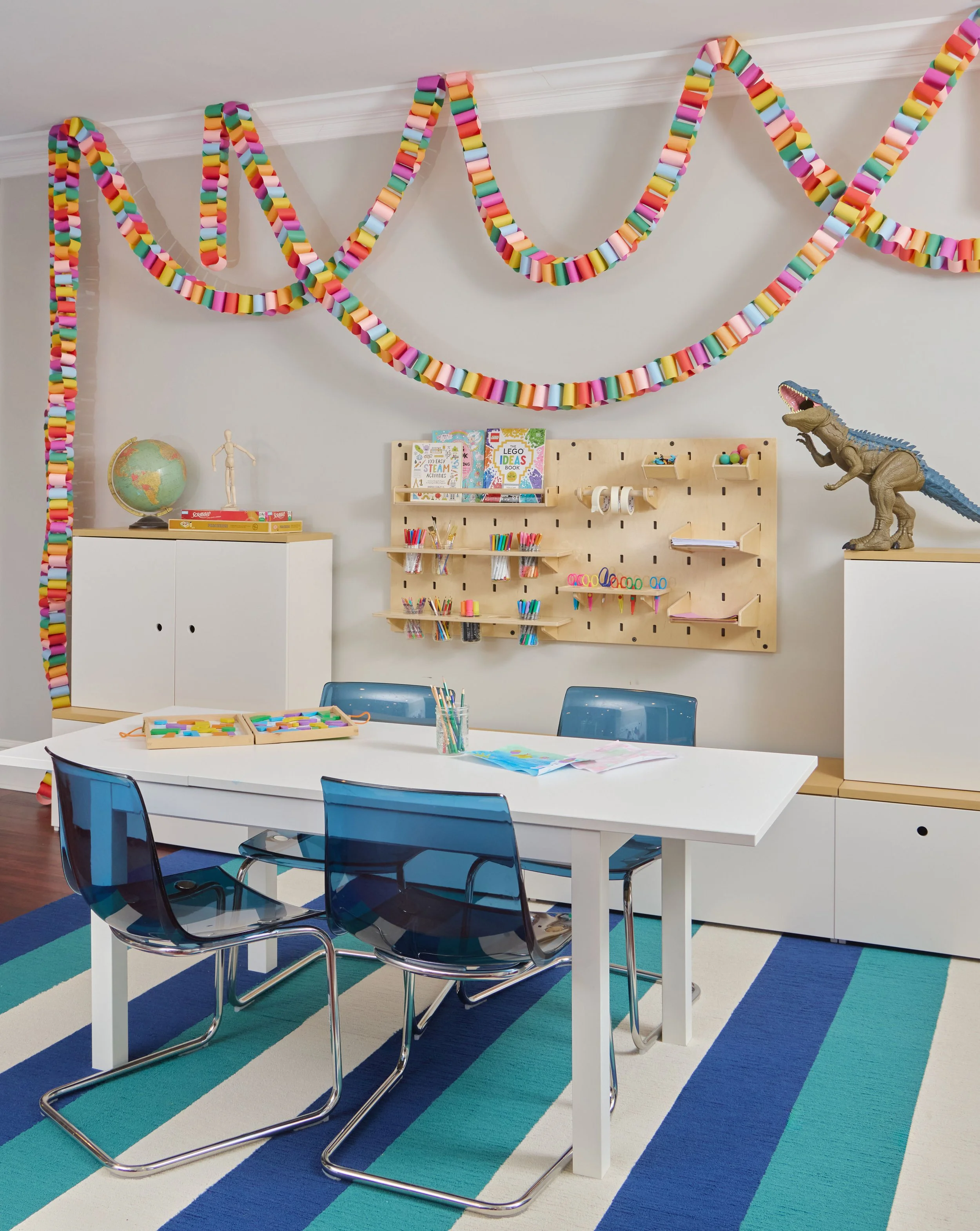 Kids' craft and homework station with white table, blue acrylic chairs, wooden pegboard shelving, and rainbow garlands