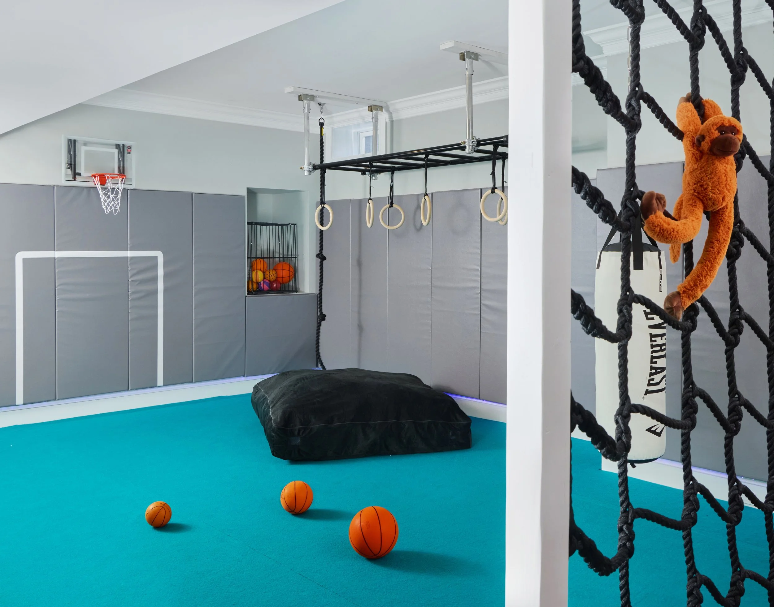 Fairfield, CT basement kids' gym with overhead monkey bars, gymnastic rings, black crash pad, and basketballs on teal turf