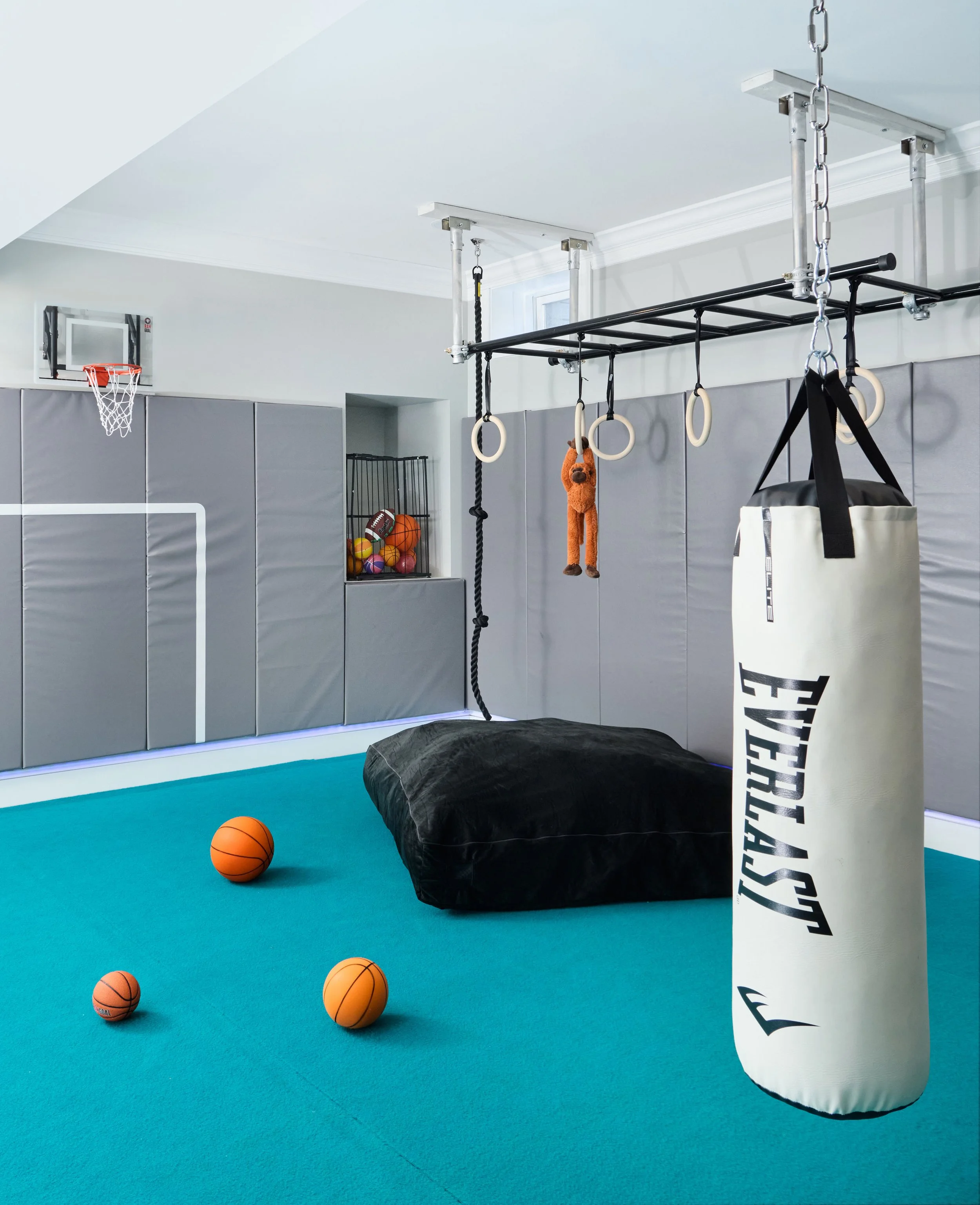 Home gym play zone with hanging punching bag, monkey bars, gymnastic rings, and mini basketball hoop over teal turf