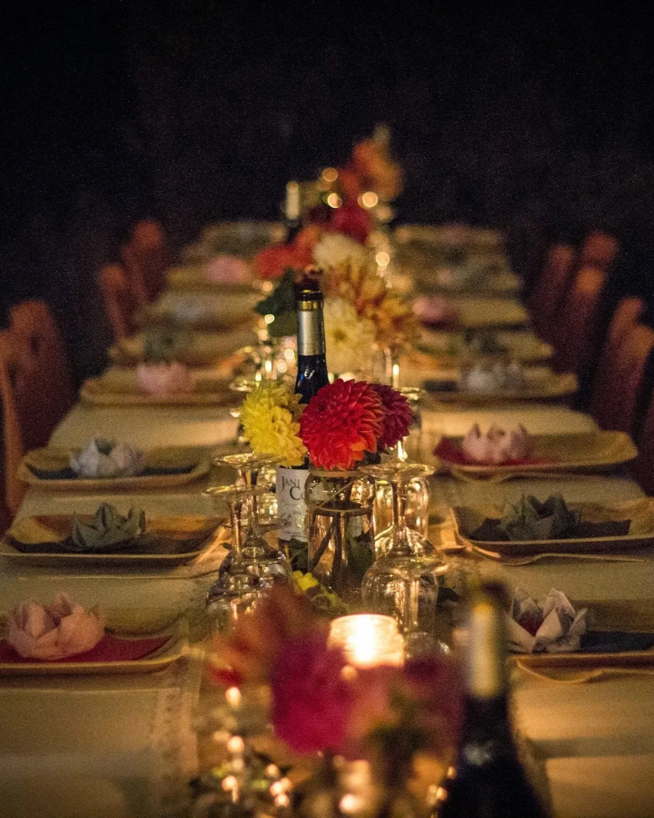 A long, elegantly set dinner table decorated with flowers, candles, and glassware at a dimly lit event.