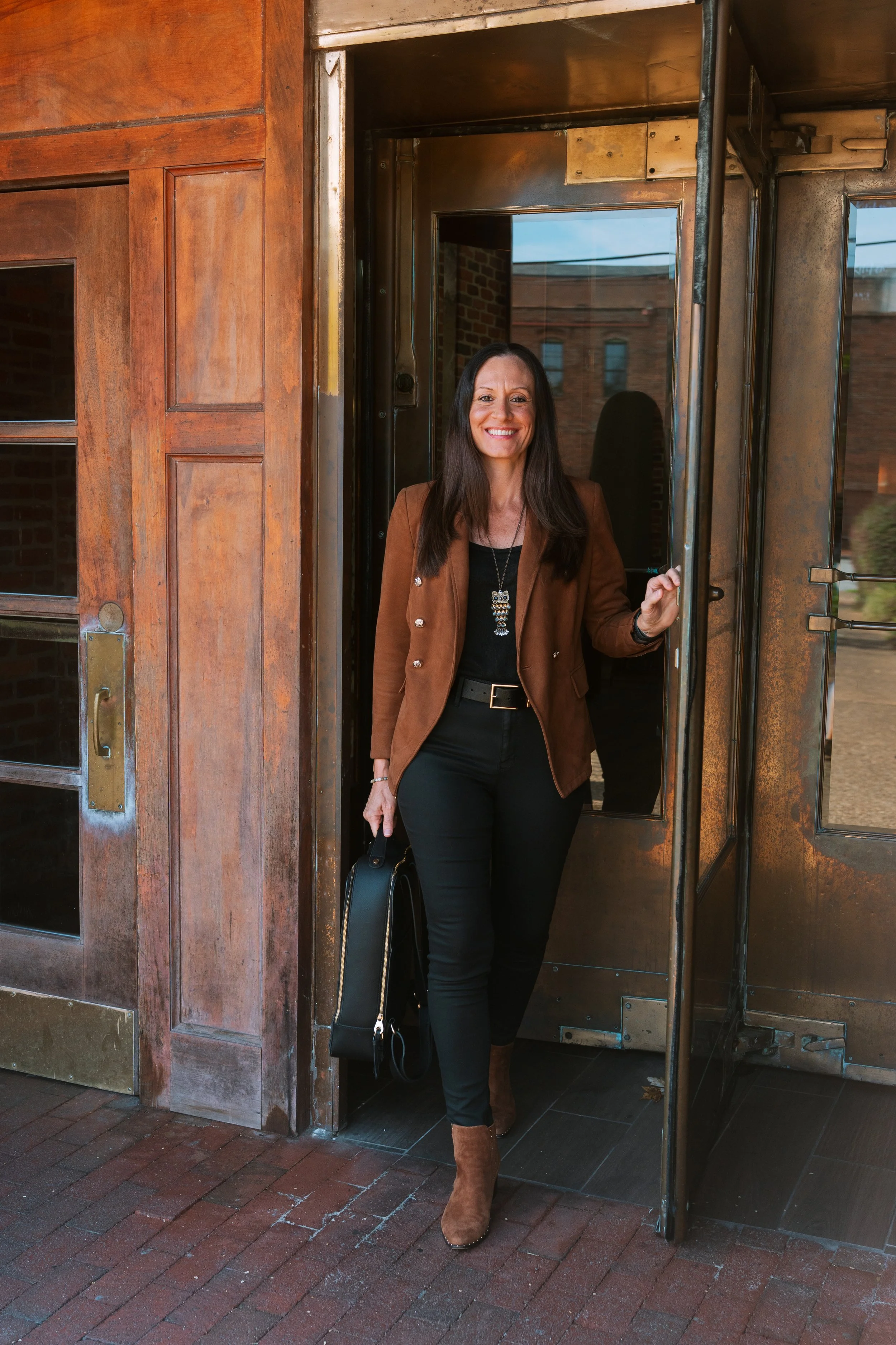 A woman with long dark hair, dressed in a brown blazer and black pants, smiling and holding a black purse, standing in the doorway of a building with a metal frame.