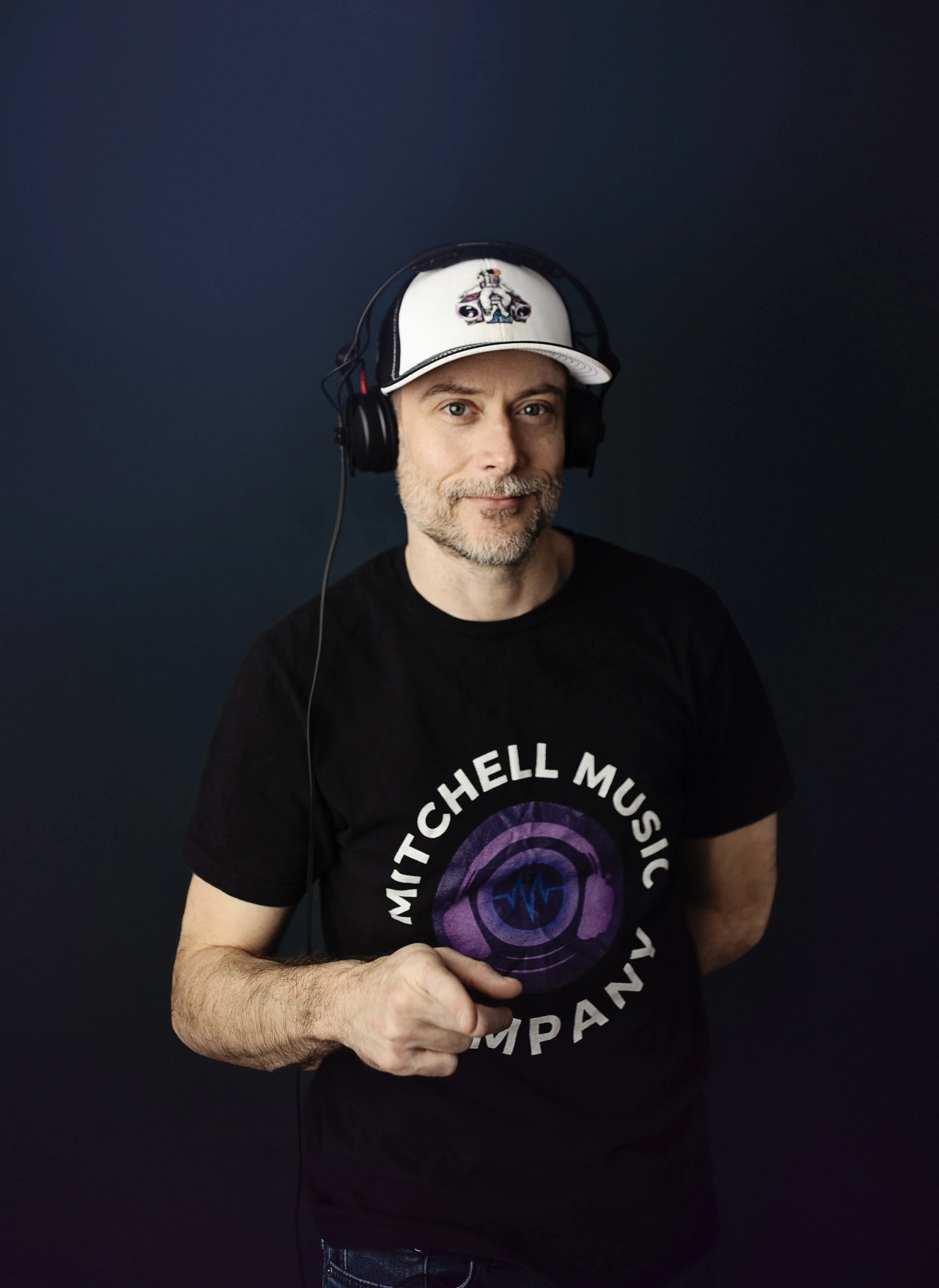 A man wearing headphones and a Mitchell Music Company T-shirt, standing against a dark background, looking at the camera with a slight smile.