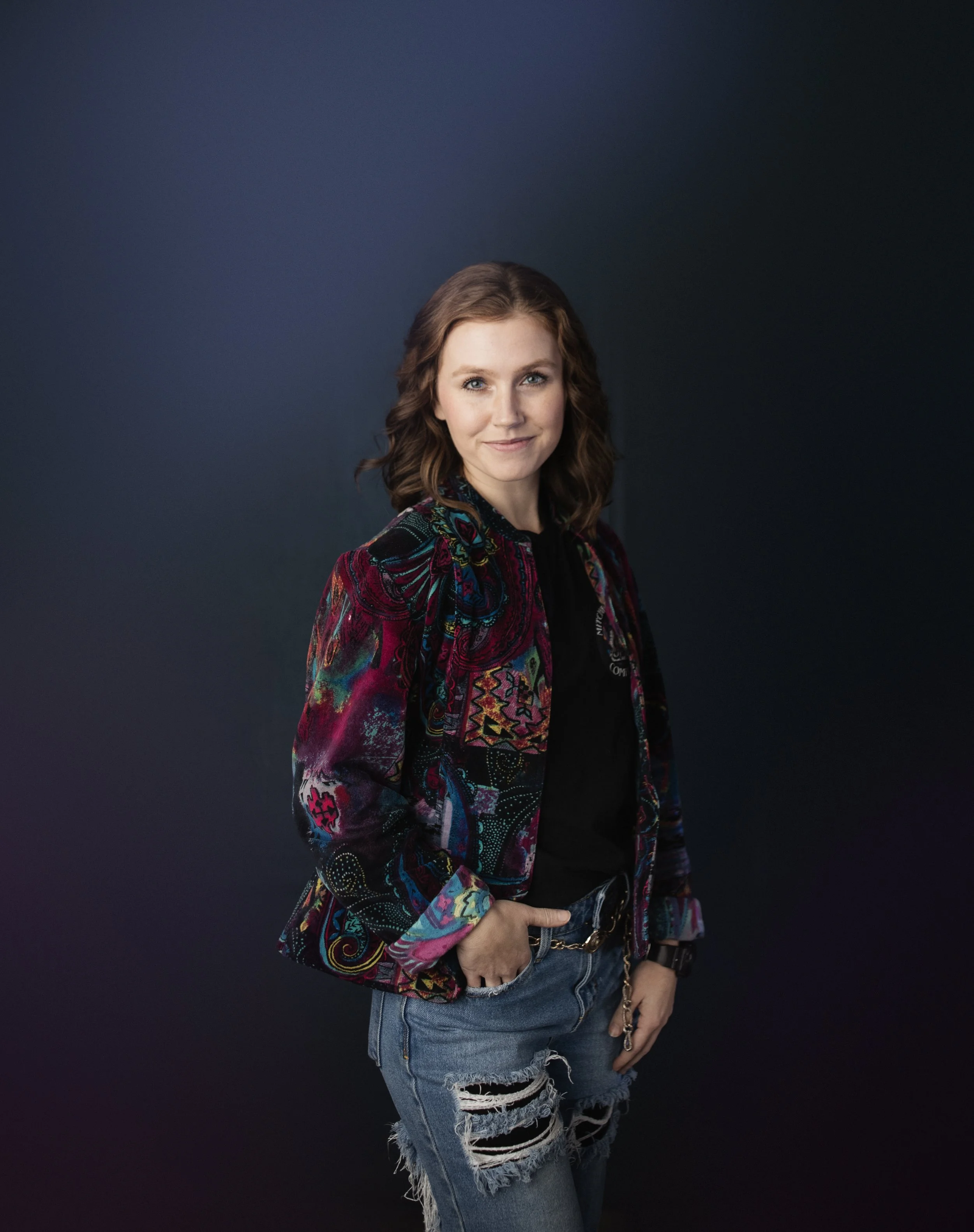A young woman with shoulder-length brown hair, wearing a colorful embroidered jacket over a black t-shirt and ripped jeans, standing against a dark background.