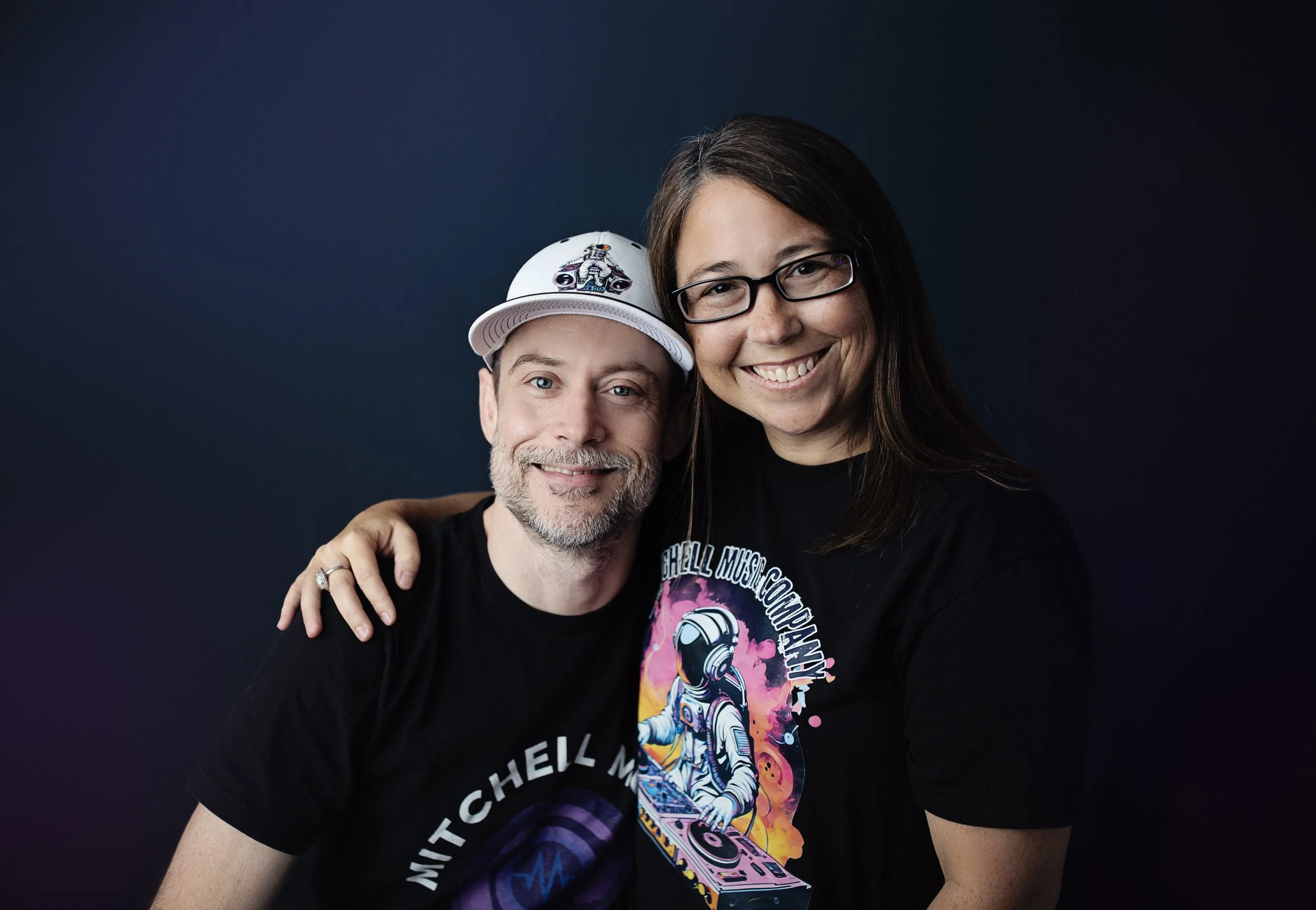 A smiling man wearing a white cap with a motorcycle design and a black T-shirt and a woman with glasses and a black T-shirt with a space-themed DJ graphic, standing close together against a dark background.