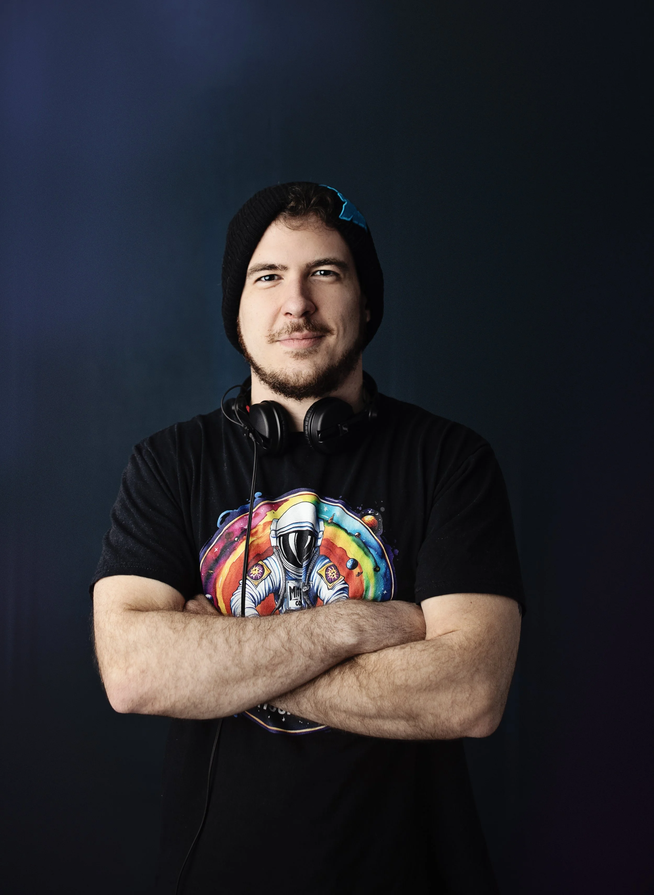 A young man with a beard and mustache wearing a black beanie, black T-shirt with colorful astronaut and rainbow design, and black headphones around his neck, standing against a dark background with arms crossed.