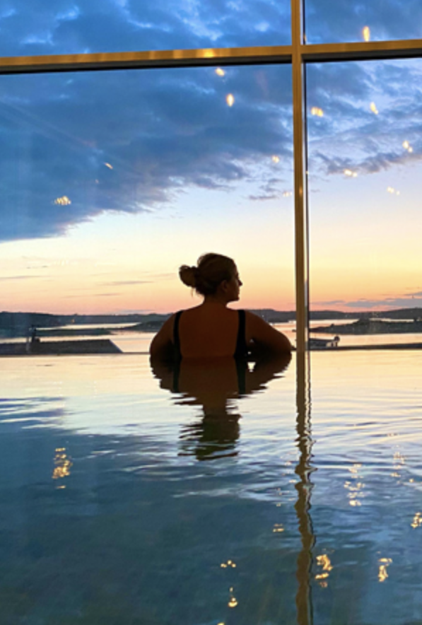 En kvinna i ett pool, som tittar ut mot en solnedgång genom ett stort fönster.