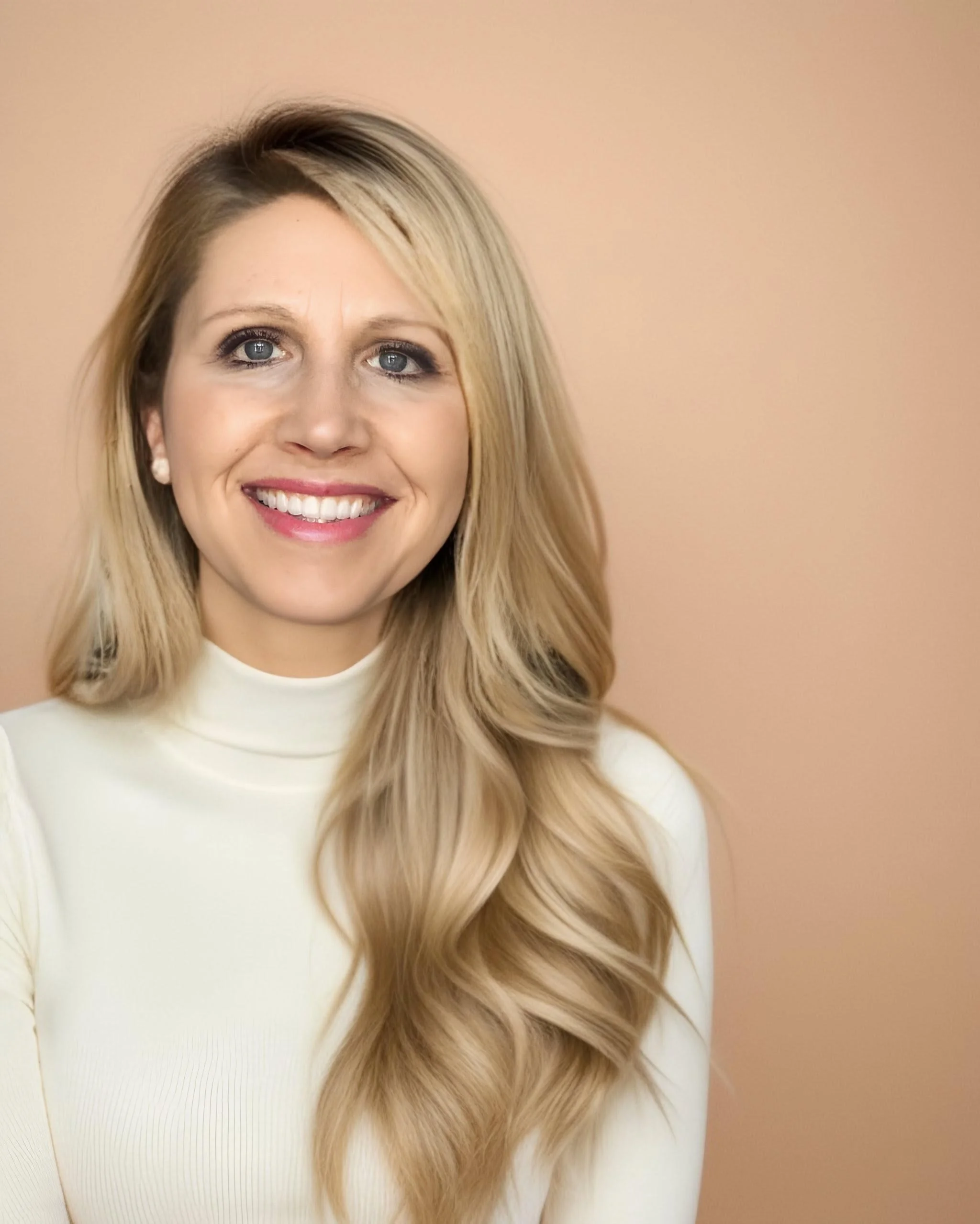 A smiling blonde woman wearing a cream-colored turtleneck shirt, with wavy hair, standing against a peach background.