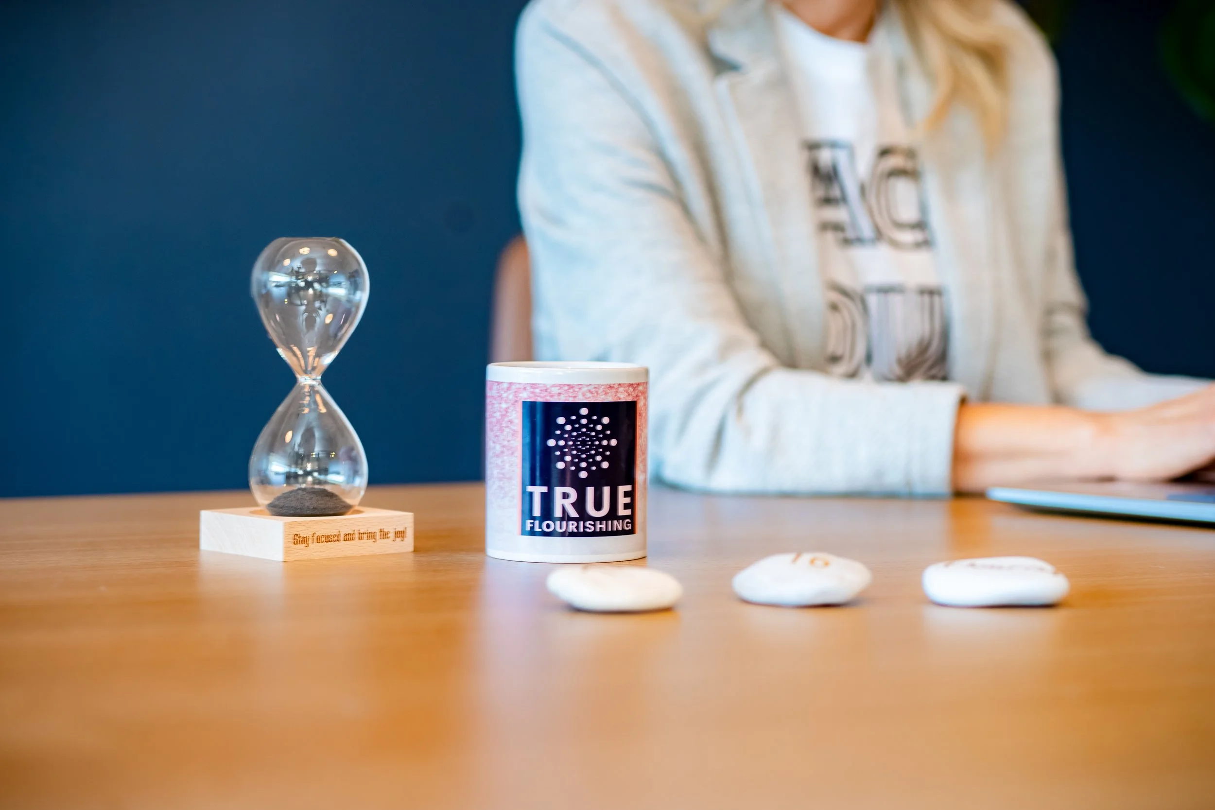 A wooden table with a clear hourglass, a mug labeled "TRUE FLOURISHING" with a dark blue and pink design, three white stones with small markings, and a person in a gray hoodie working on a laptop in the background.