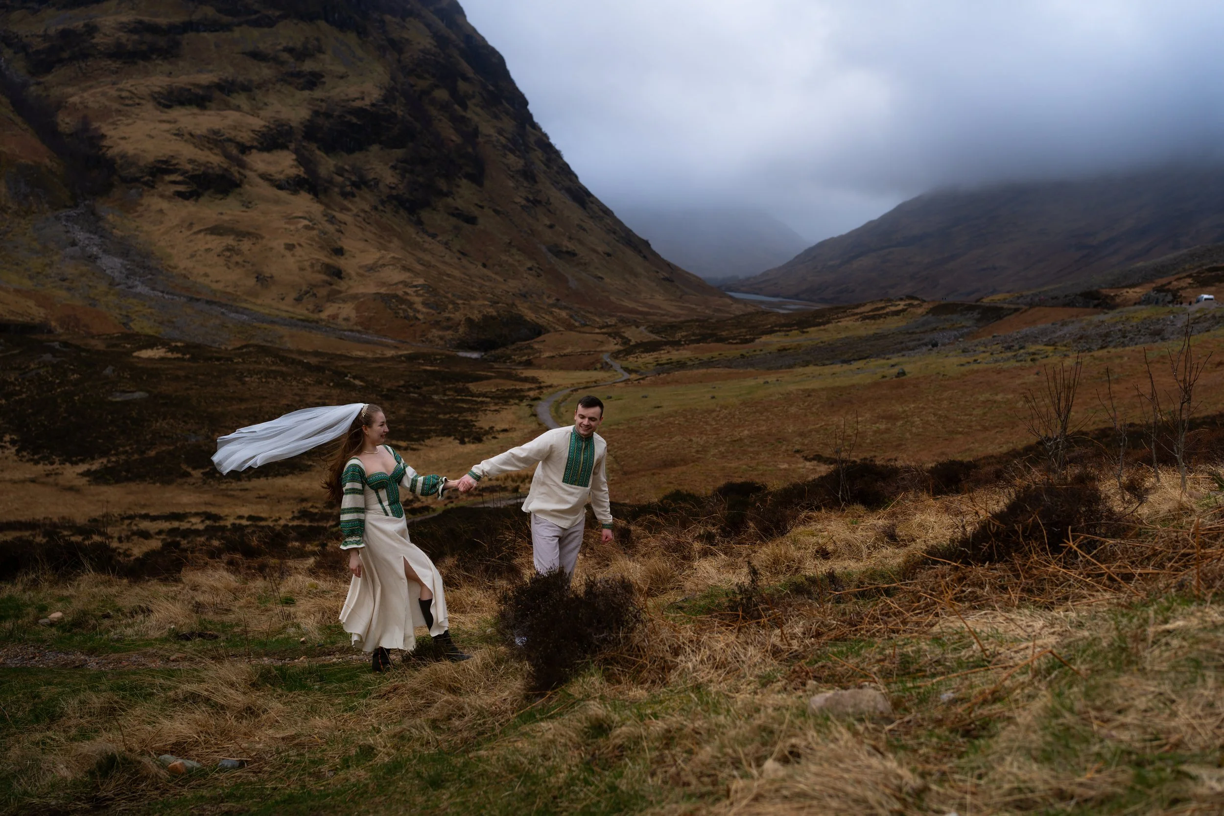 A couple in traditional clothing holding hands and walking in a rugged, hilly landscape with mountains in the background and overcast skies.
