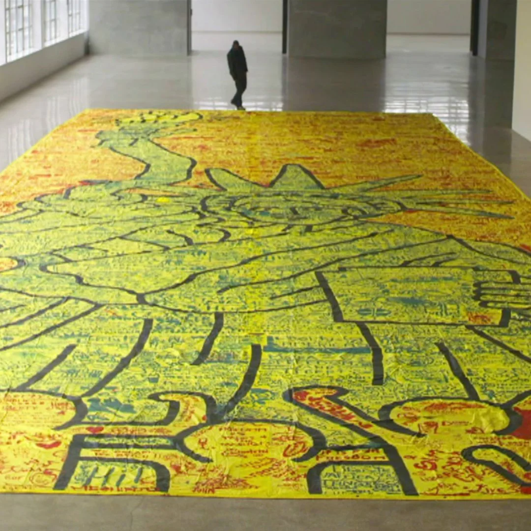 Large colorful artwork on the floor of a gallery, featuring a predominantly yellow background with black outline drawings of a dog, with some areas filled with red and blue markings.