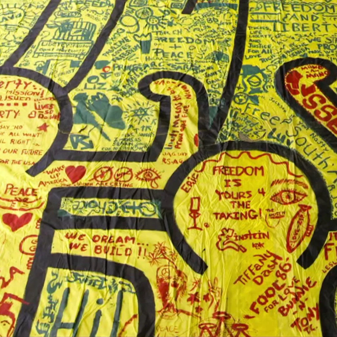 Close-up of a yellow cloth with numerous handwritten messages, quotes, and drawings in red, green, blue, and black ink. Prominent words include 'FREEDOM,' 'PEACE,' and 'LIBERTY.' The messages advocate for freedom, peace, and justice, with various symbols and personal notes.