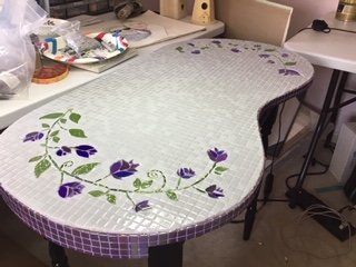 A curved table with a decorative mosaic surface featuring purple flower patterns and green leaves, surrounded by a cluttered workspace.