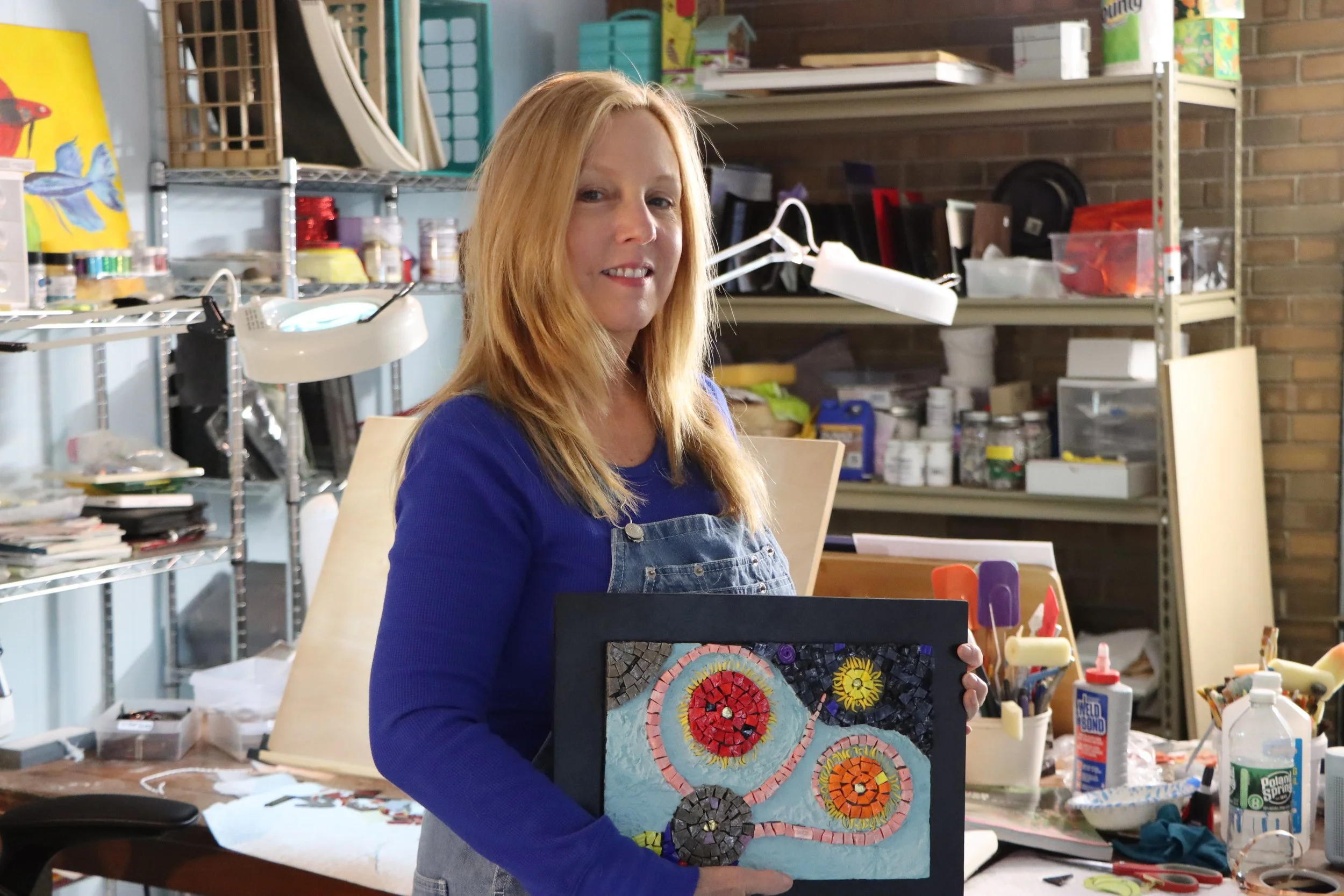 A woman with long red hair wearing a blue top and denim apron holding a colorful mosaic art piece in a cluttered art studio.