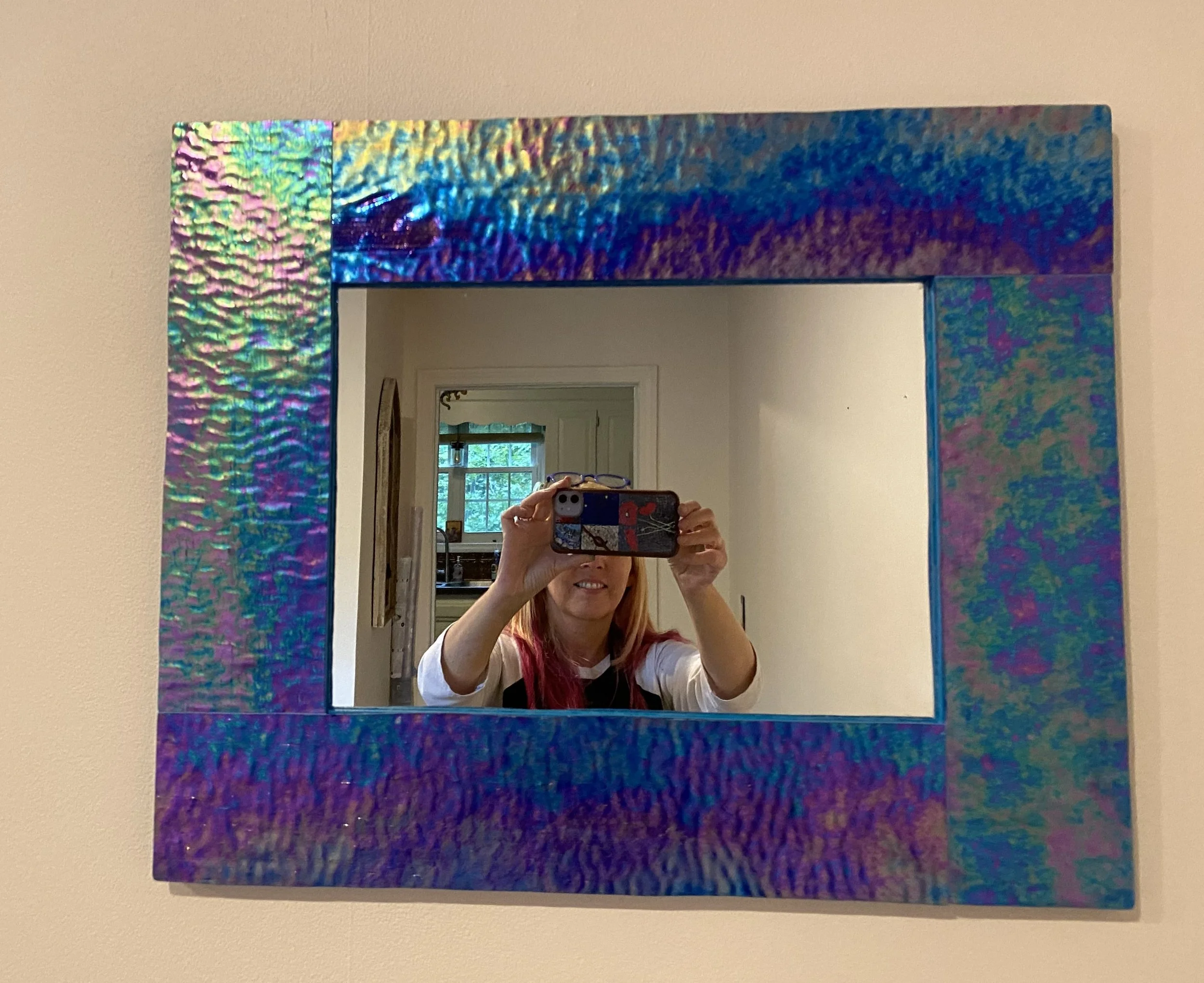 Person taking a selfie in front of a colorful, reflective square mirror with a textured frame, standing in a room with a window and kitchen in the background.