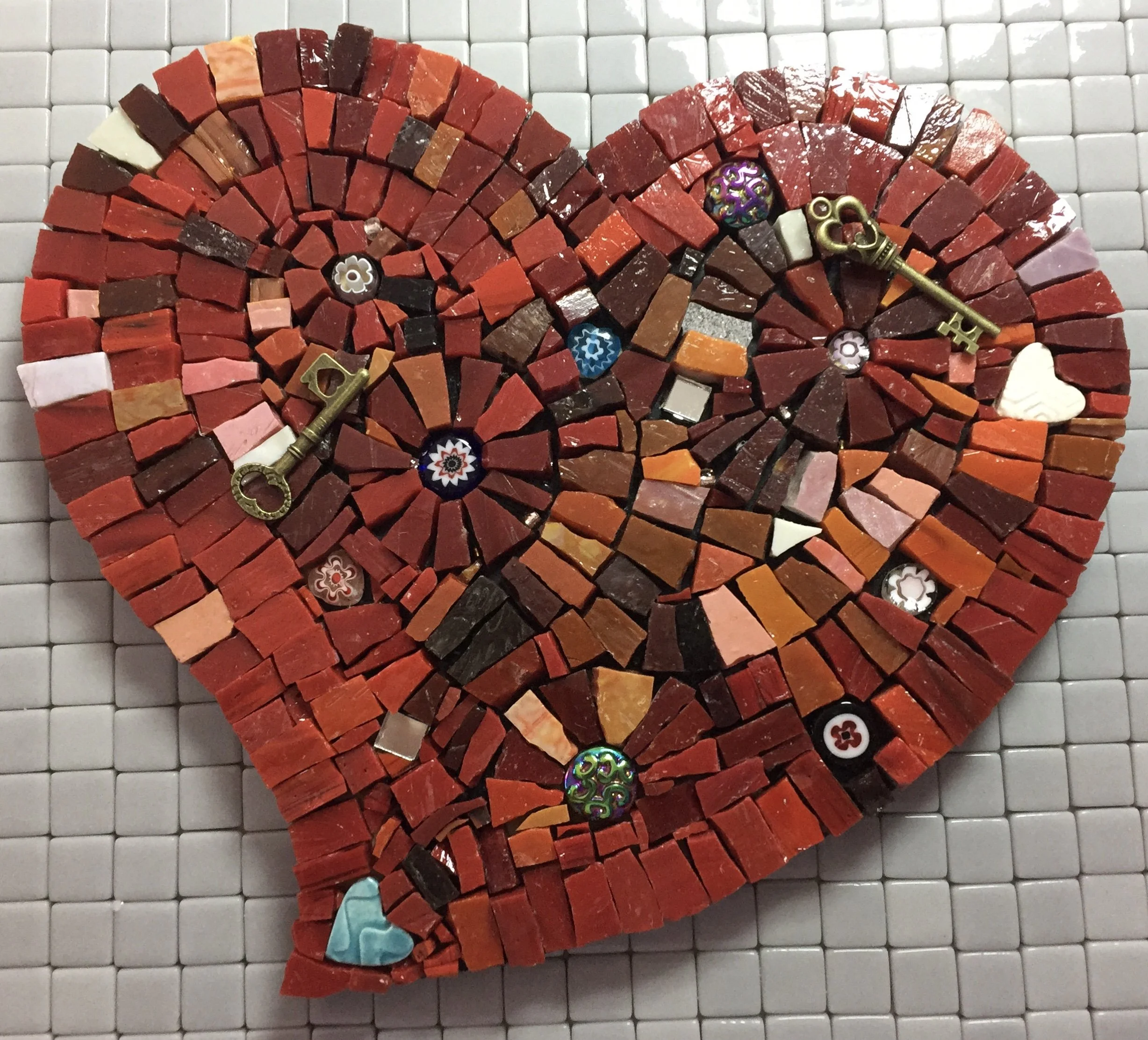 Red mosaic heart made of small stone tiles with decorative keys and buttons, placed on a white tiled background.
