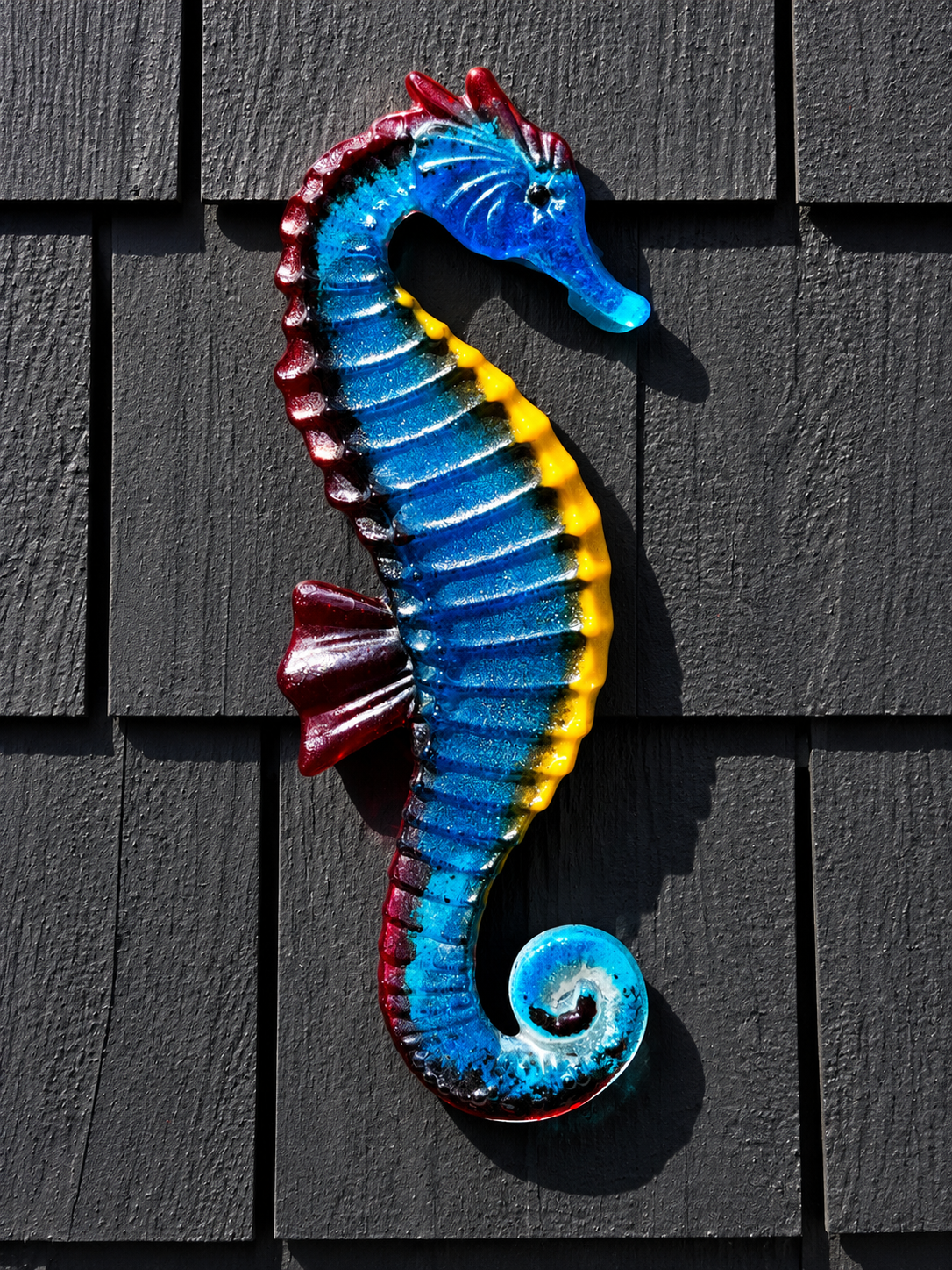 Handmade blue fused glass seahorse wall art with turquoise, yellow and red accents displayed on a dark exterior wall