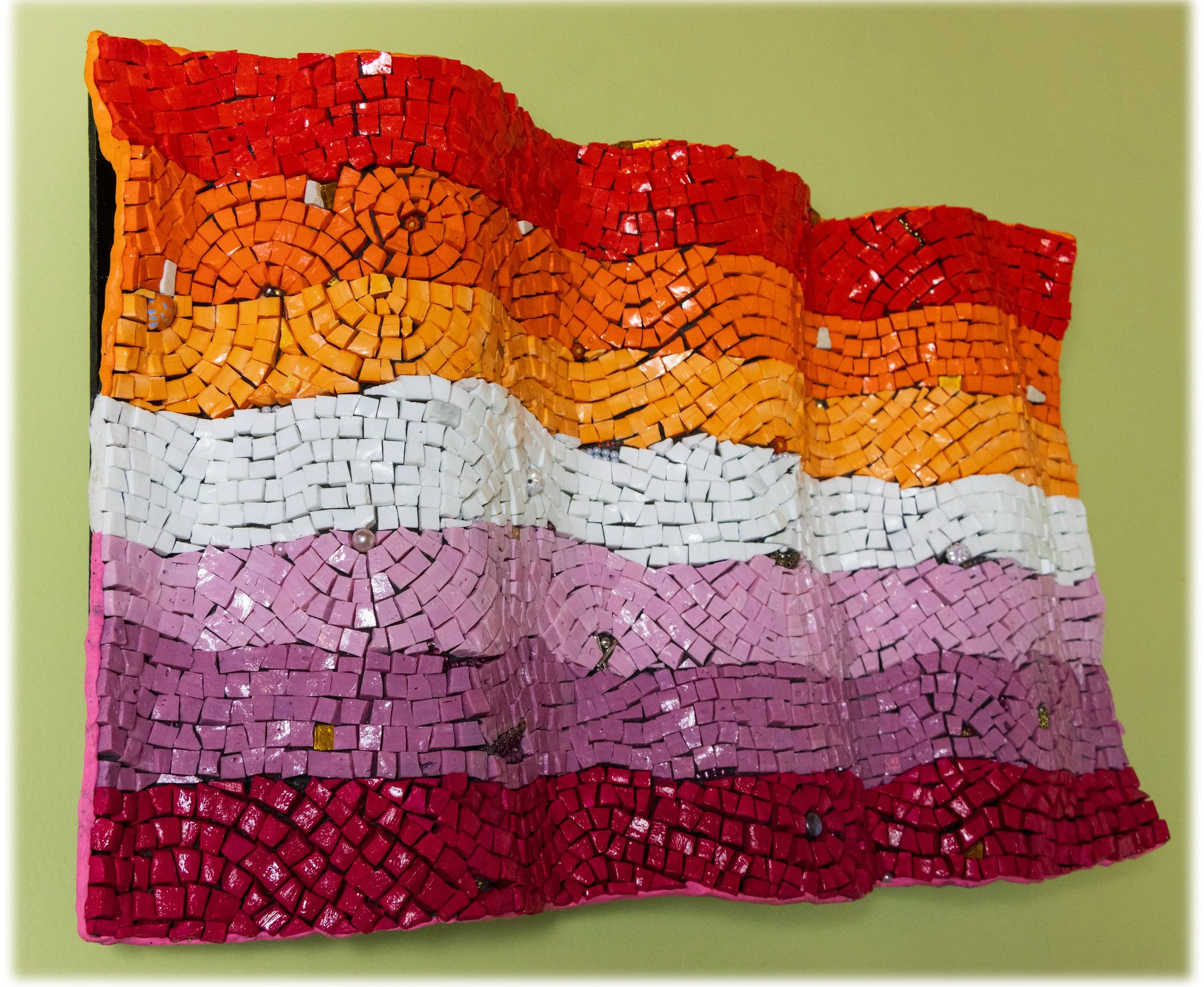 Colorful mosaic artwork made of small, square tiles arranged in wavy stripes of red, orange, white, pink, purple, and red on a light green background.