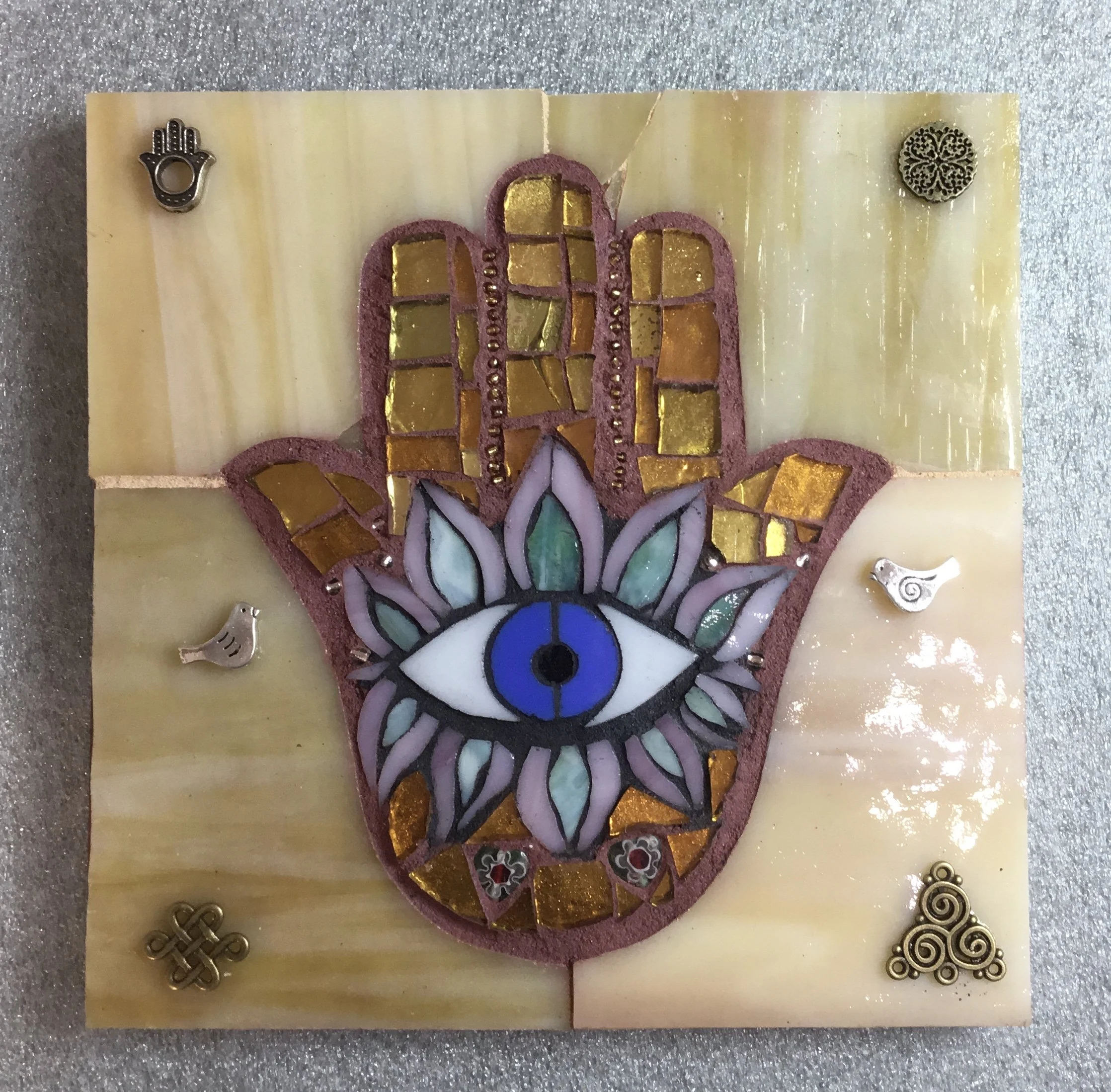 Decorative mosaic art featuring a Hamsa hand with an eye in the center, surrounded by colorful glass and metal embellishments on a marble background.