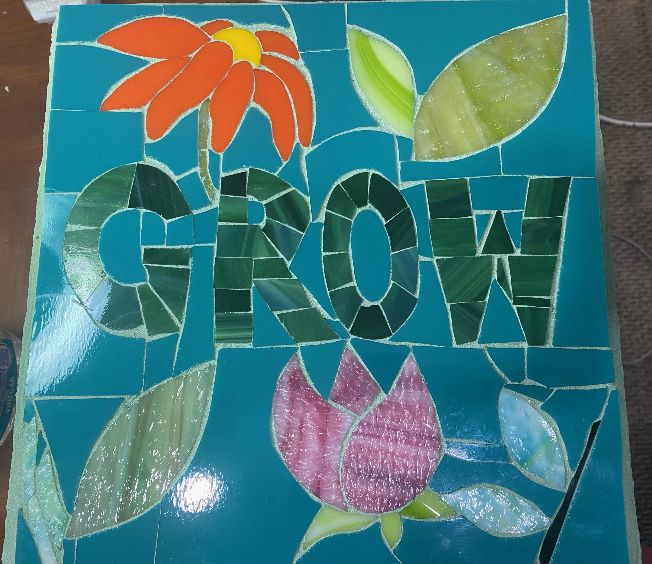 A colorful mosaic with the phrase 'LOVE' in large letters, surrounded by flowers, leaves, and abstract shapes, all made of stained glass tiles on a blue background.