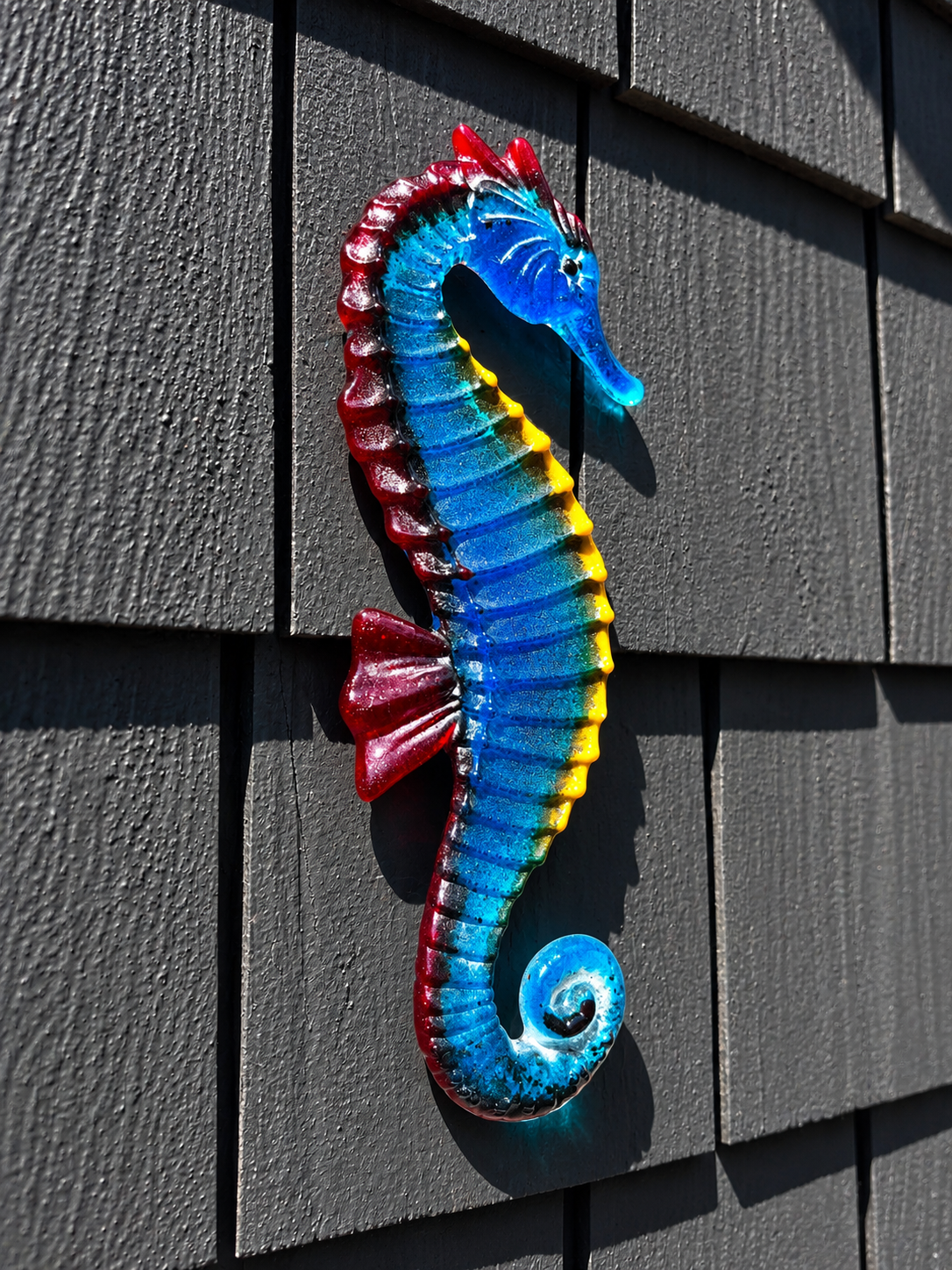 Handmade blue fused glass seahorse wall art with turquoise, yellow and red accents displayed on a dark exterior wall
