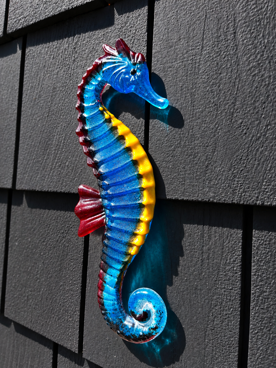 Handmade blue fused glass seahorse wall art with turquoise, yellow and red accents displayed on a dark exterior wall