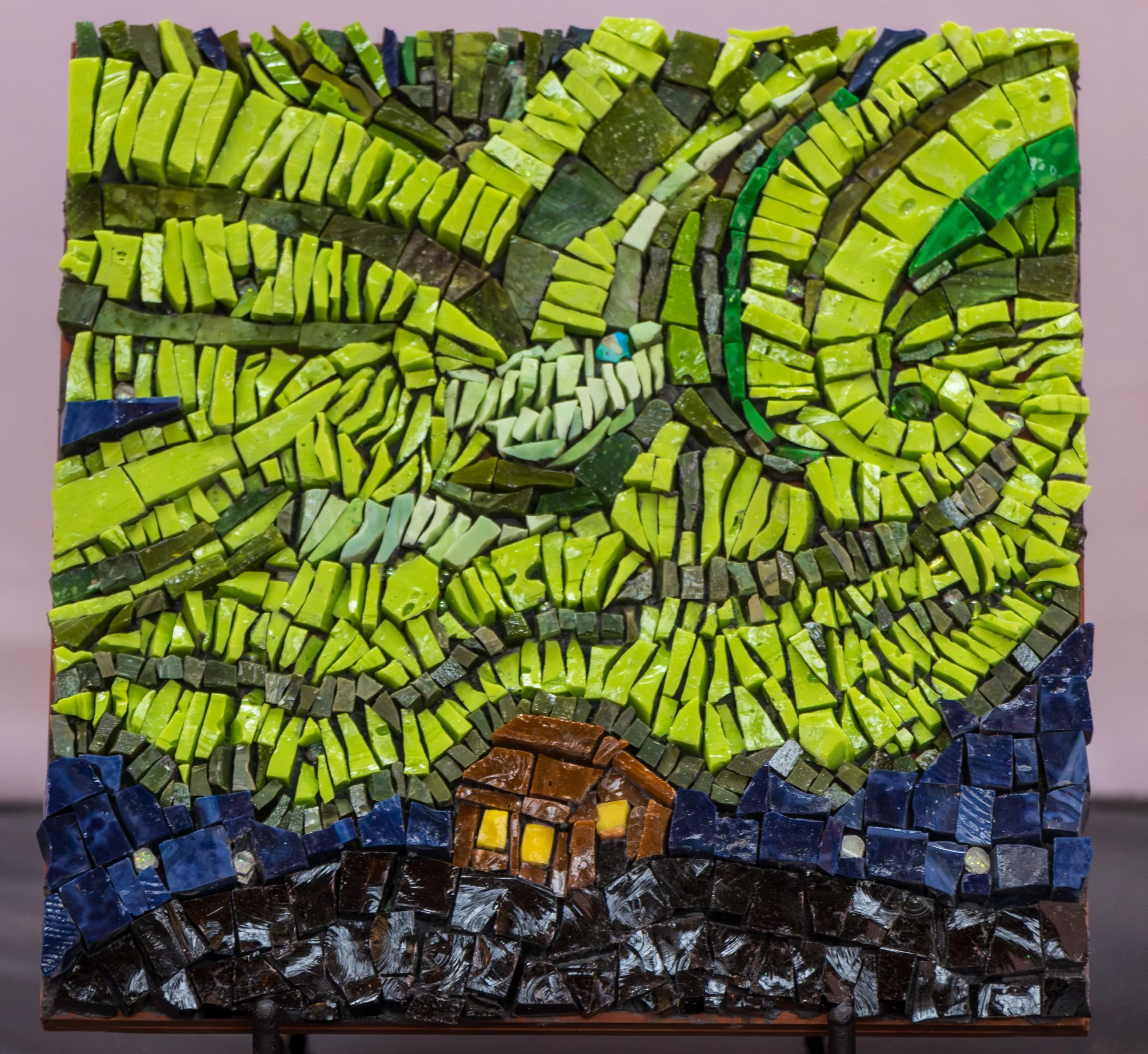 Mosaic artwork depicting a colorful landscape scene with green rolling hills, a small yellow house, and a blue sky made from small, irregularly shaped glass pieces.
