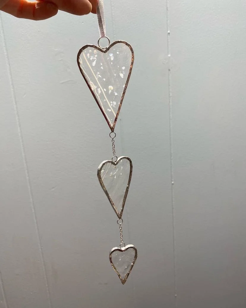 Hand holding a hanging decoration with three clear glass hearts connected by small metal links against a plain background.