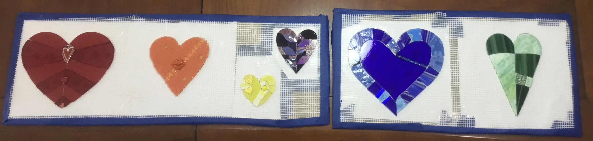 Two blue-framed display boards with various colorful mosaic hearts made of different materials, arranged in a line on a wooden surface.
