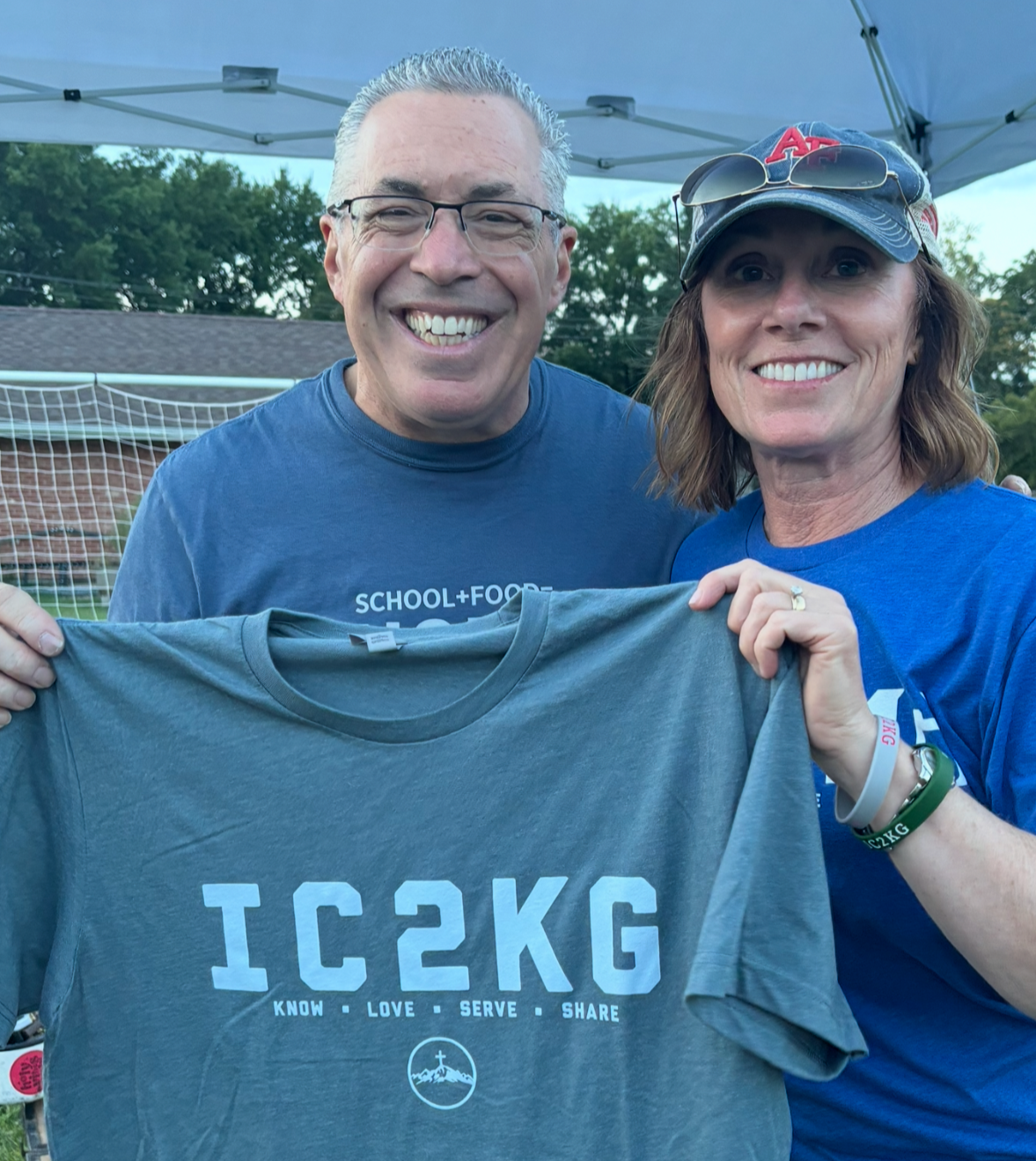 A smiling man and woman holding a gray t-shirt that has 'IC 2KG' and the words 'Know, Love, Serve, Share' printed on it, standing outdoors under a tent.