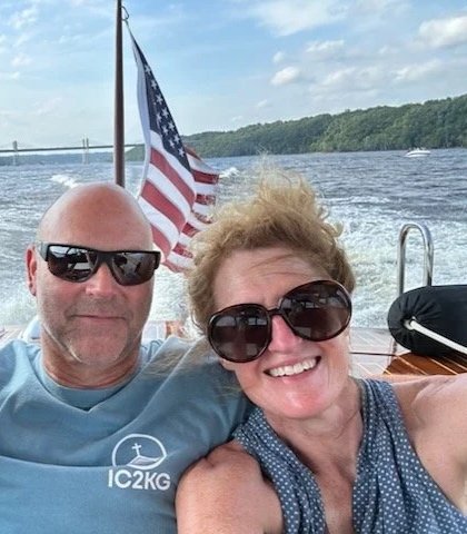 Three smiling people, two women and one man, taking a selfie on a boat with an American flag in the background on a body of water with a bridge and green trees in the distance.