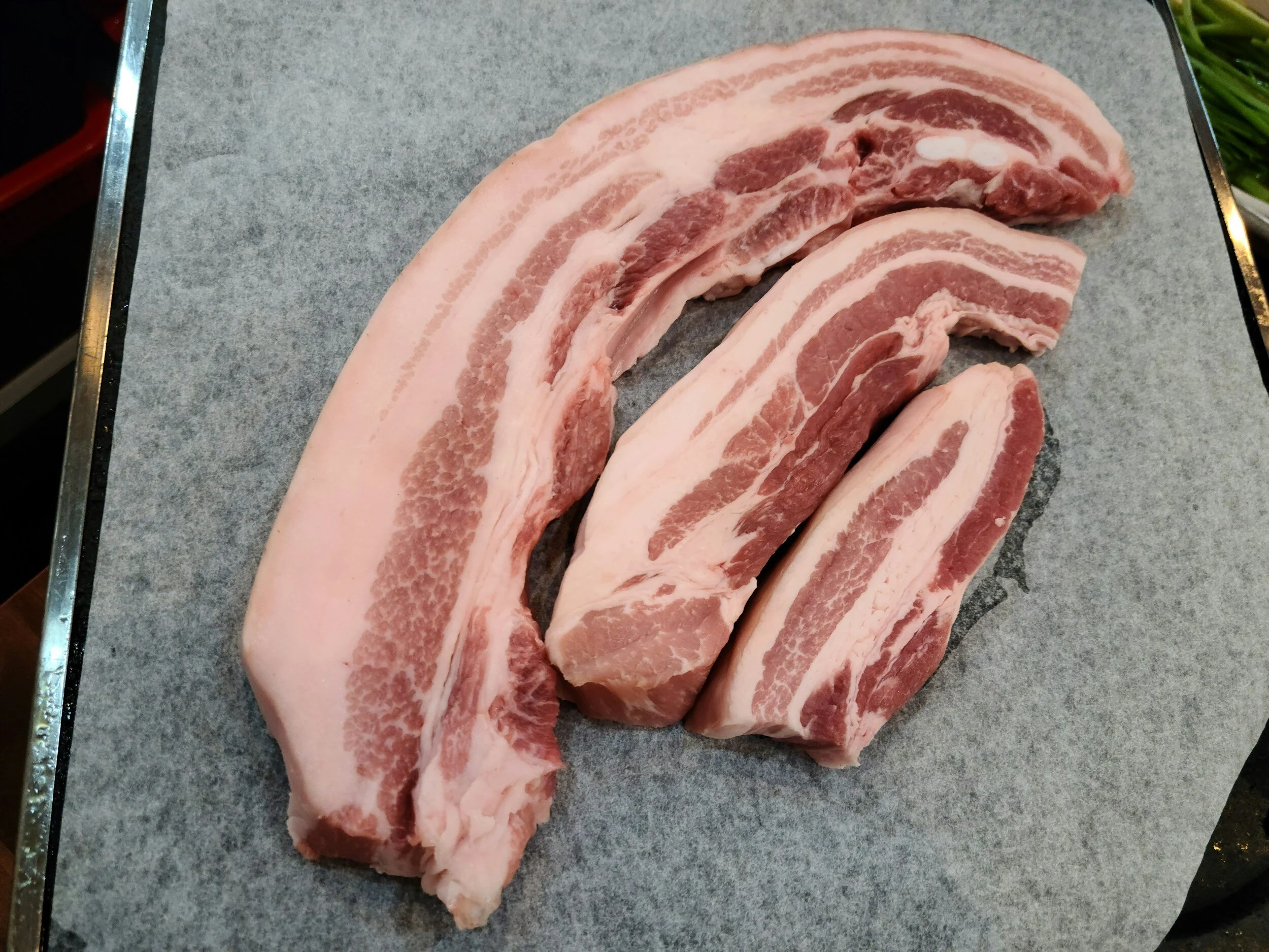 Slices of raw pastured pork belly with layers of meat and fat on a piece of gray parchment paper.