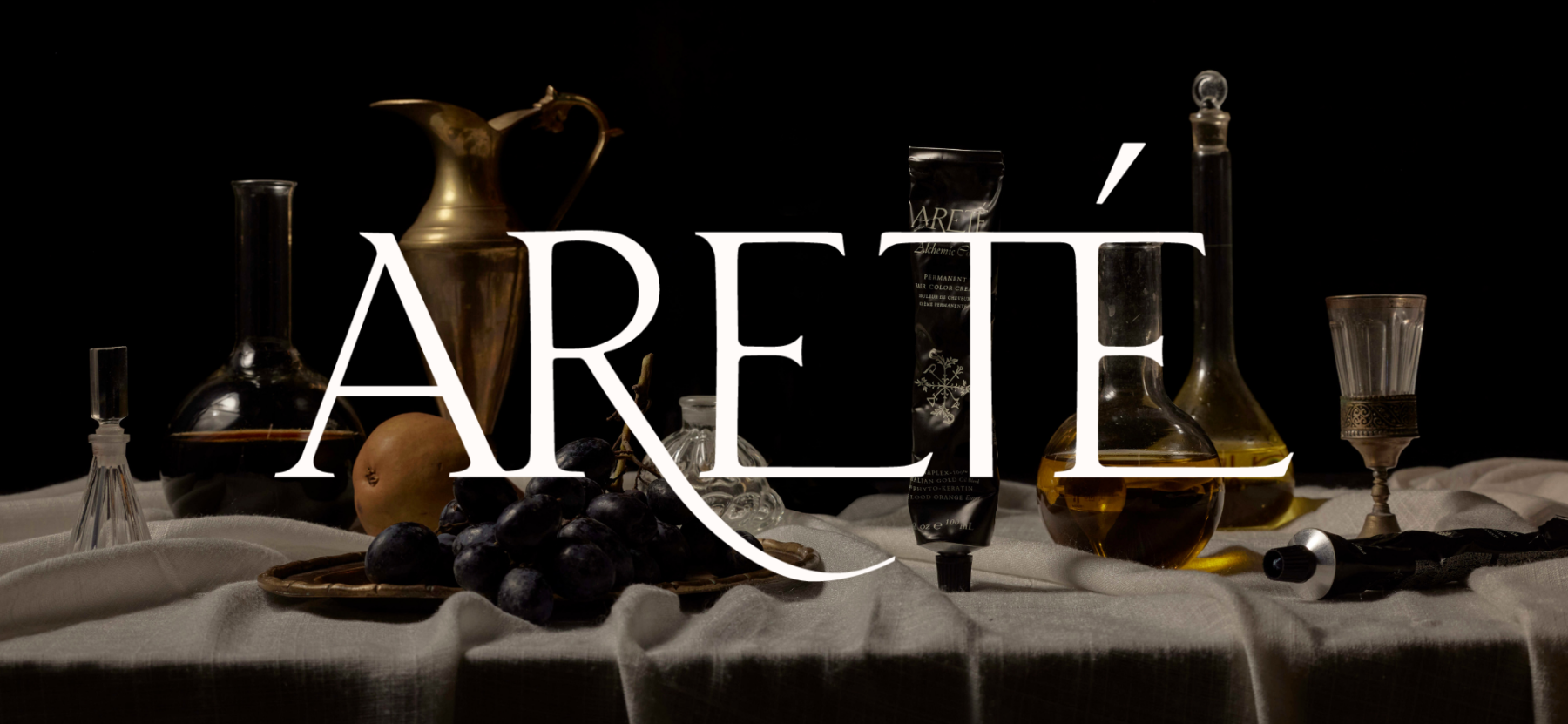 Still life arrangement of antique glass bottles, grapes, a pear, and various cosmetic or perfume items on a white cloth, with the text 'ARÉITÉ' overlaying the scene.
