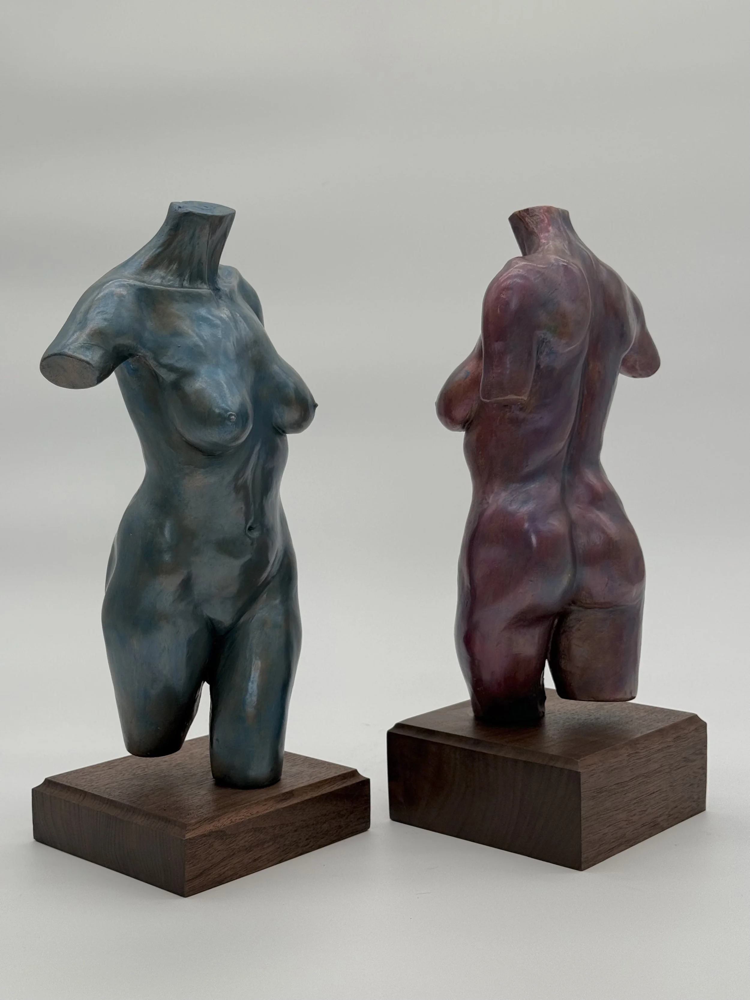 Two female torso sculptures on wooden pedestals, one in bluish hues and the other in reddish tones.