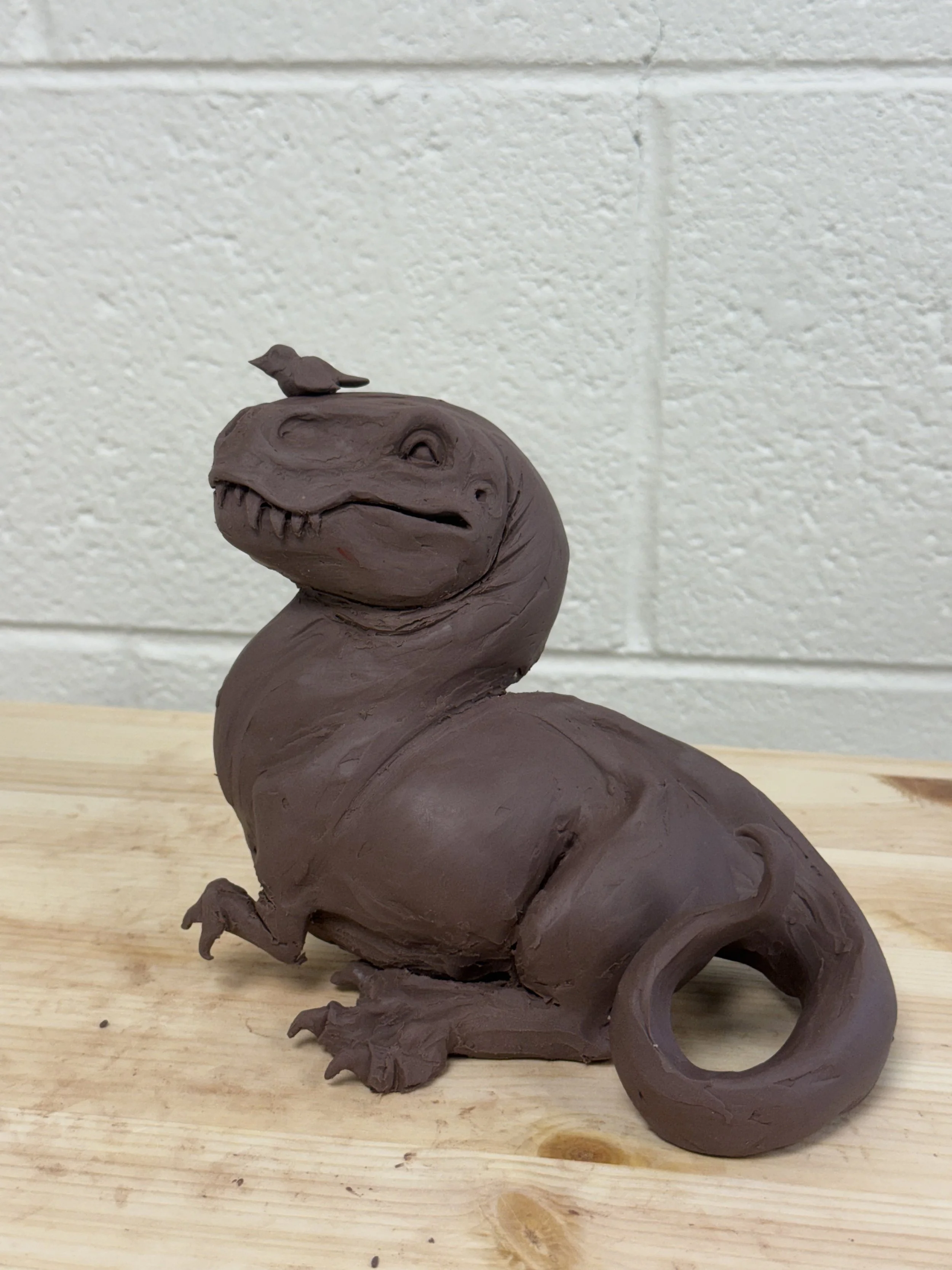 A clay sculpture of a dinosaur with a small bird on its head, placed on a wooden surface against a white brick wall background.
