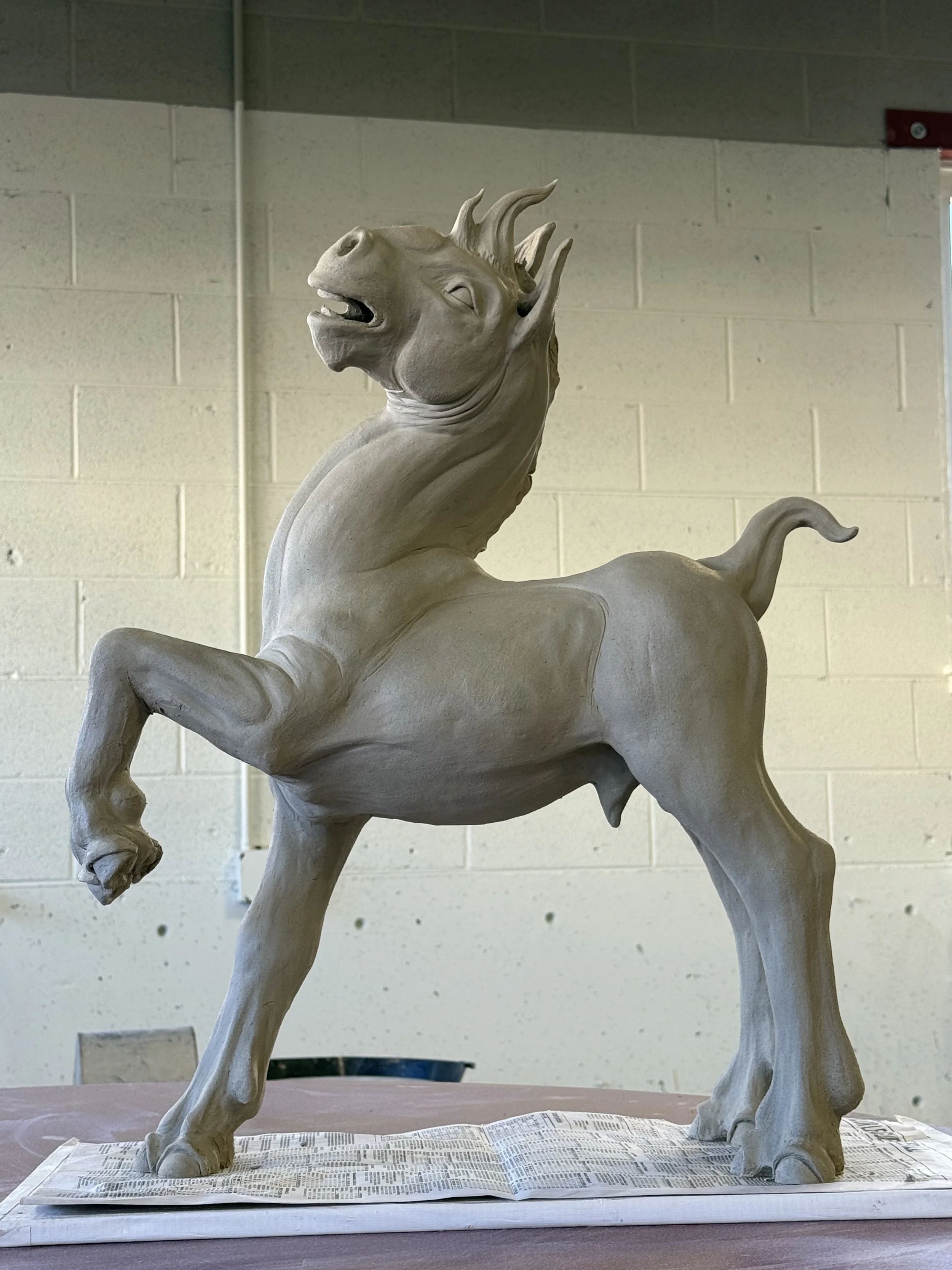 A clay sculpture of a mythical creature with a horse-like body, a lion-like head, and a mane or crest, displayed indoors on a table with newspaper.