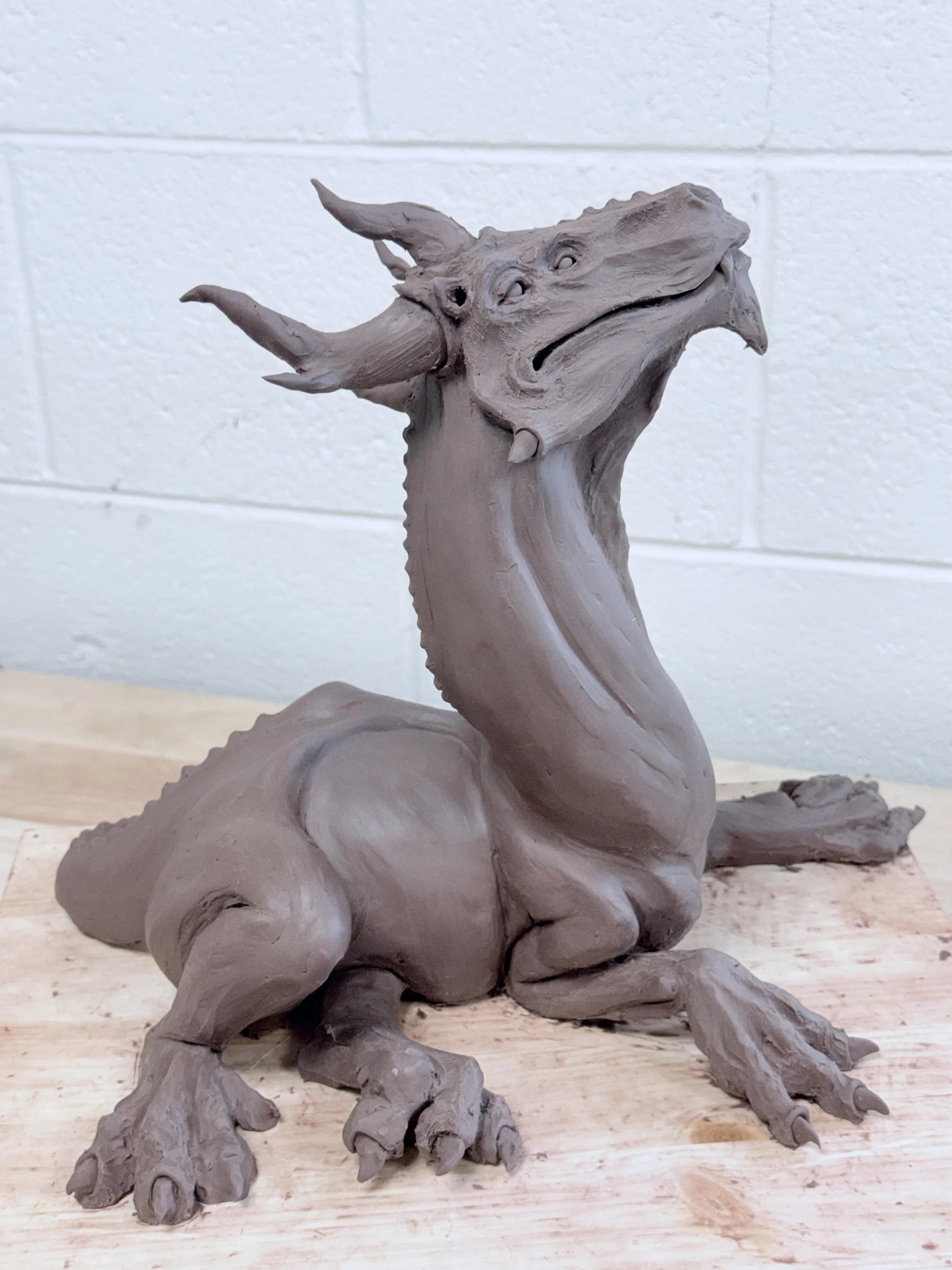 A clay sculpture of a dragon with horns, a long neck, wings, and detailed claws, sitting on a wooden surface against a white brick wall background.