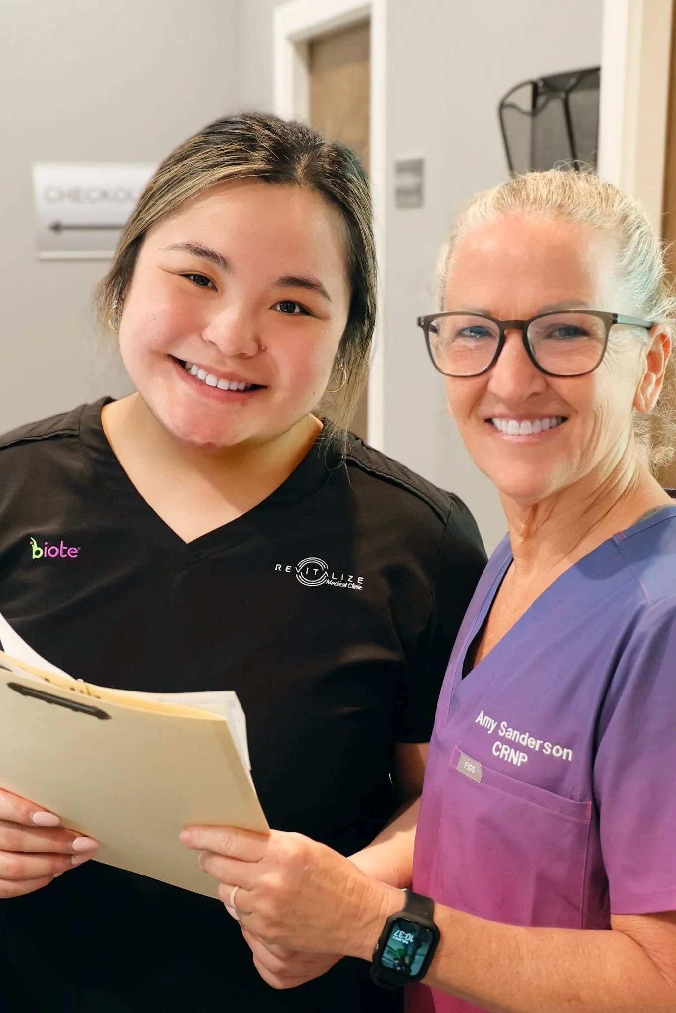 Two women, one in medical scrubs and the other in black scrubs, smiling for a selfie in a medical setting.