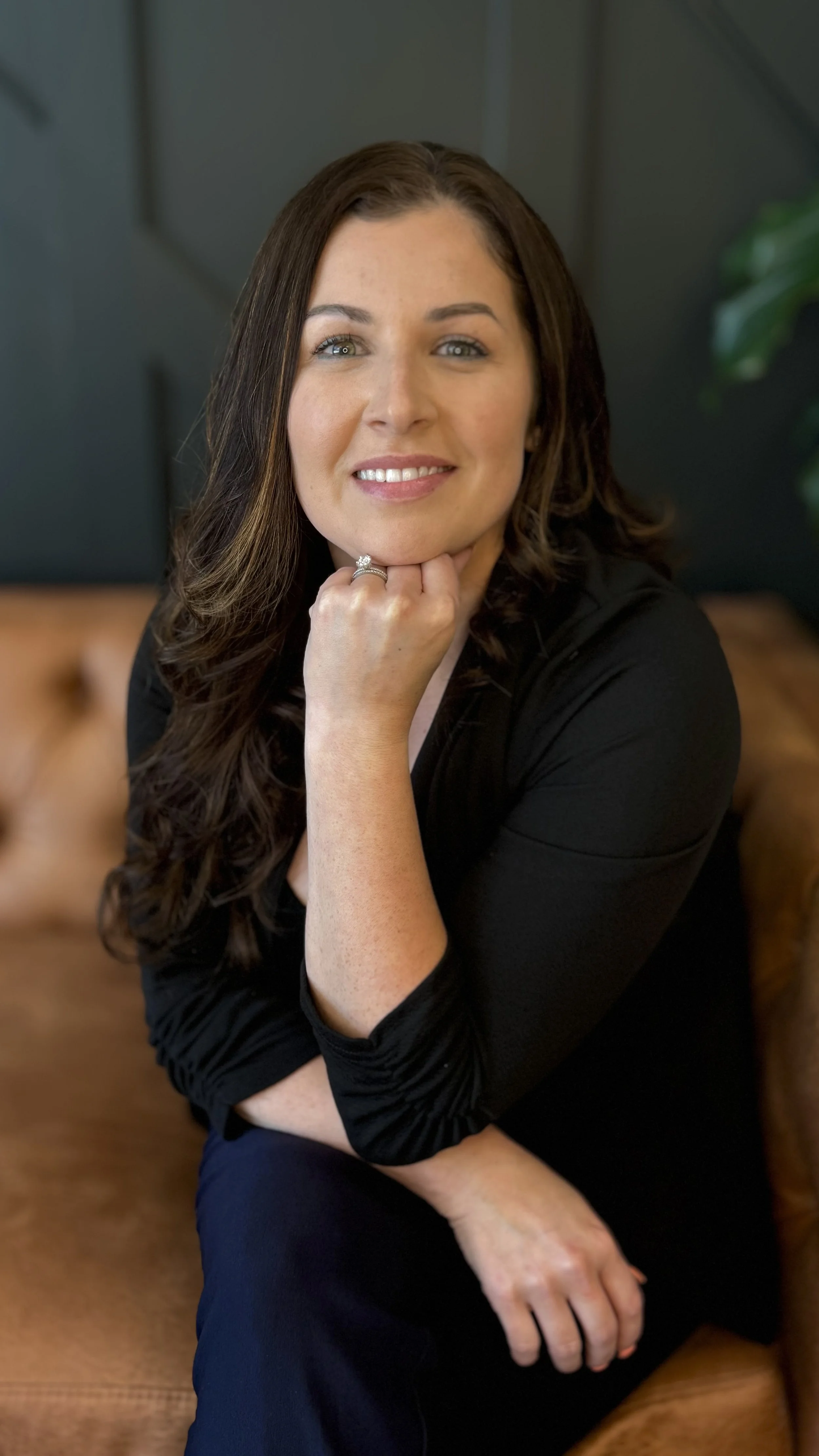 A woman with long brown hair smiling and resting her chin on her hand, sitting on a brown leather couch, wearing a black top and a ring on her finger.