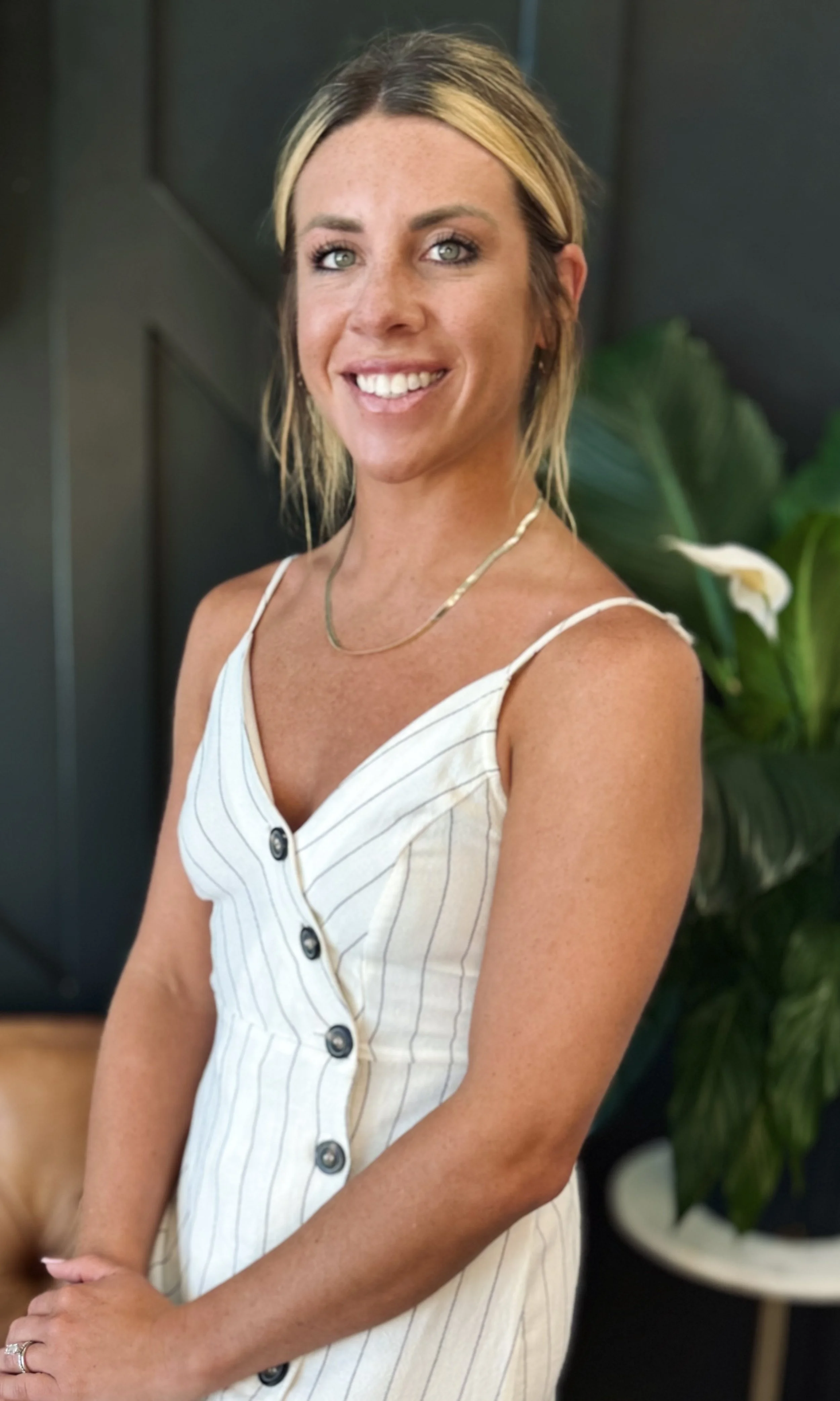 A woman with blonde hair styled with dark roots and a smile, wearing a white, buttoned, sleeveless dress with thin straps and a gold necklace, standing in front of a dark wall with a large green leafy plant in the background.