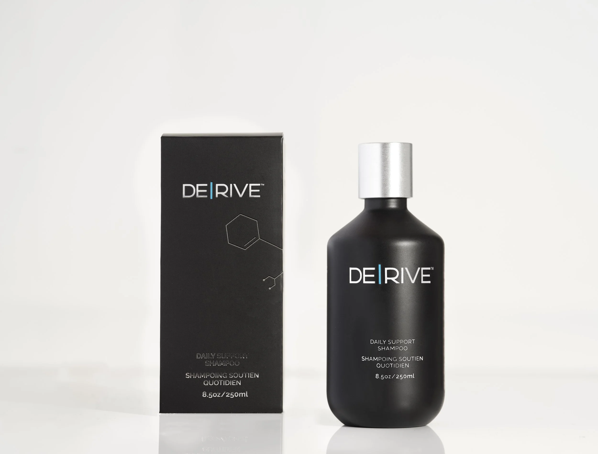 Black bottle of DE|RIVE Daily Support Shampoo with silver cap next to a black box with the same branding, set against a white background.