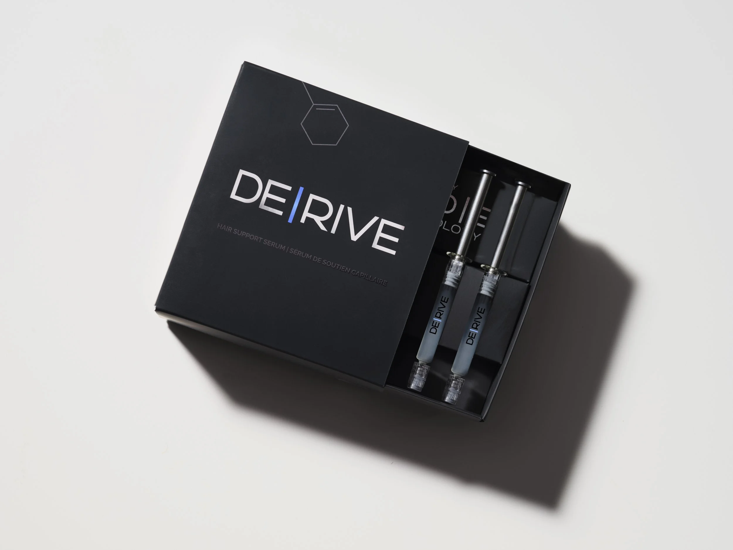 A black box labeled 'DE|DRIVE' contains two syringes filled with a clear liquid, designed for hair support treatment.