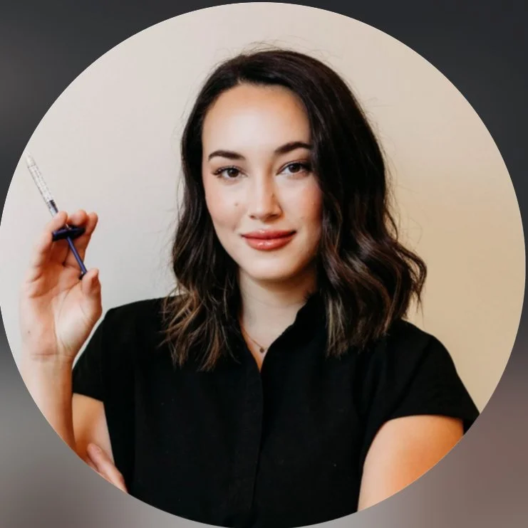 A woman with shoulder-length dark wavy hair wearing a black shirt, holding a syringe, and smiling.